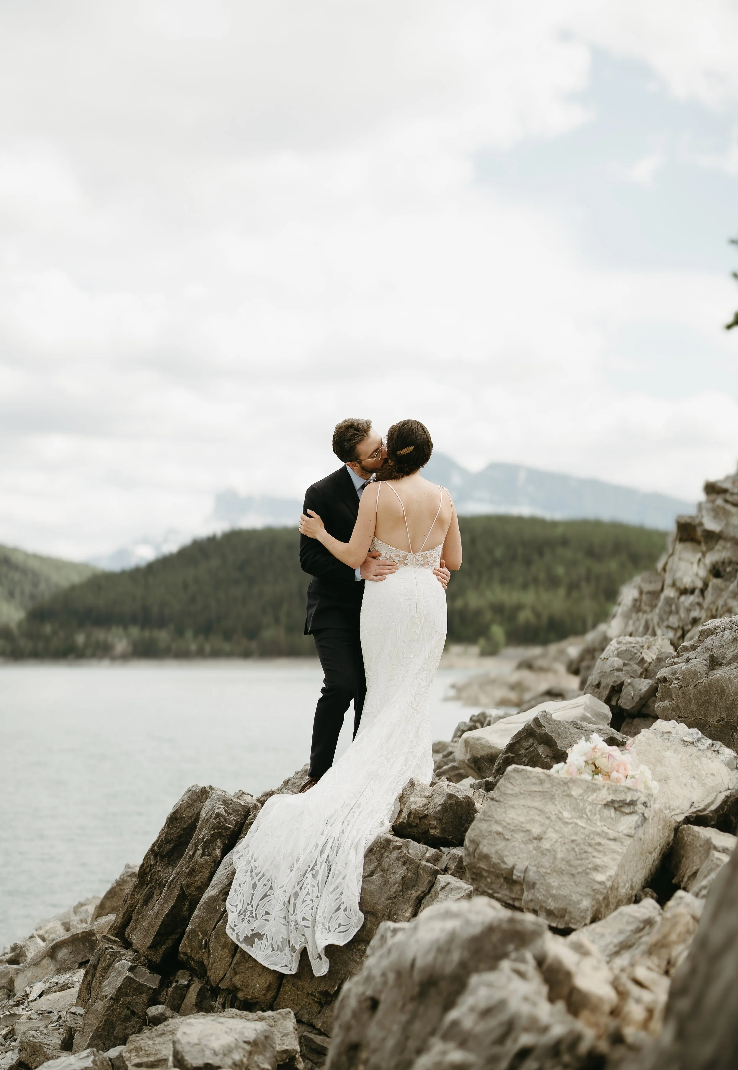 A bride and groom share a kiss on rocks beside a lake surrounded by mountains. July 2025 Banff Elopement/Wedding Session with KC Photography