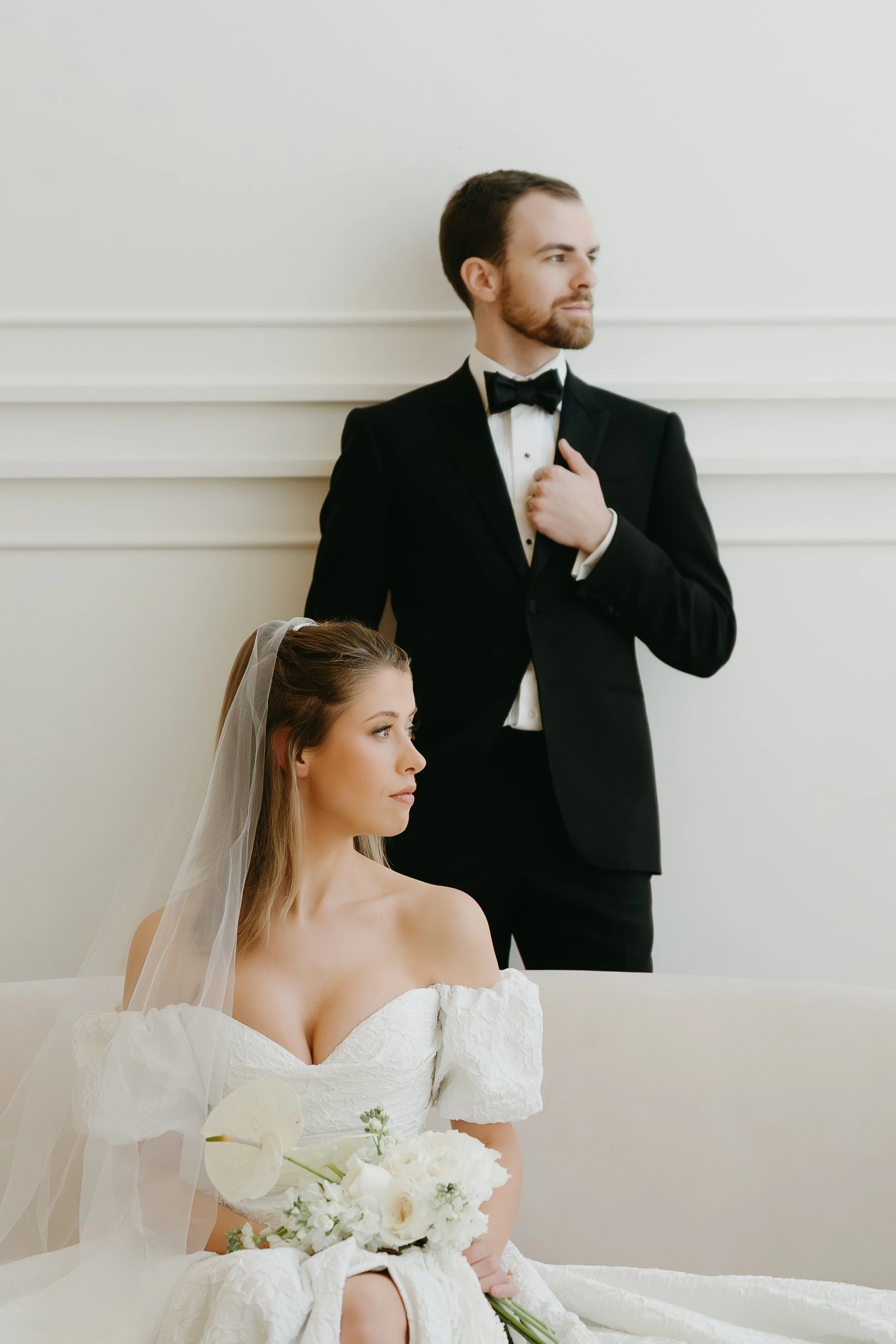 A bride sits on a couch as her groom stands behind her and they look off into the distance. Calgary Elopement/Wedding Editorial Session with KC Photography