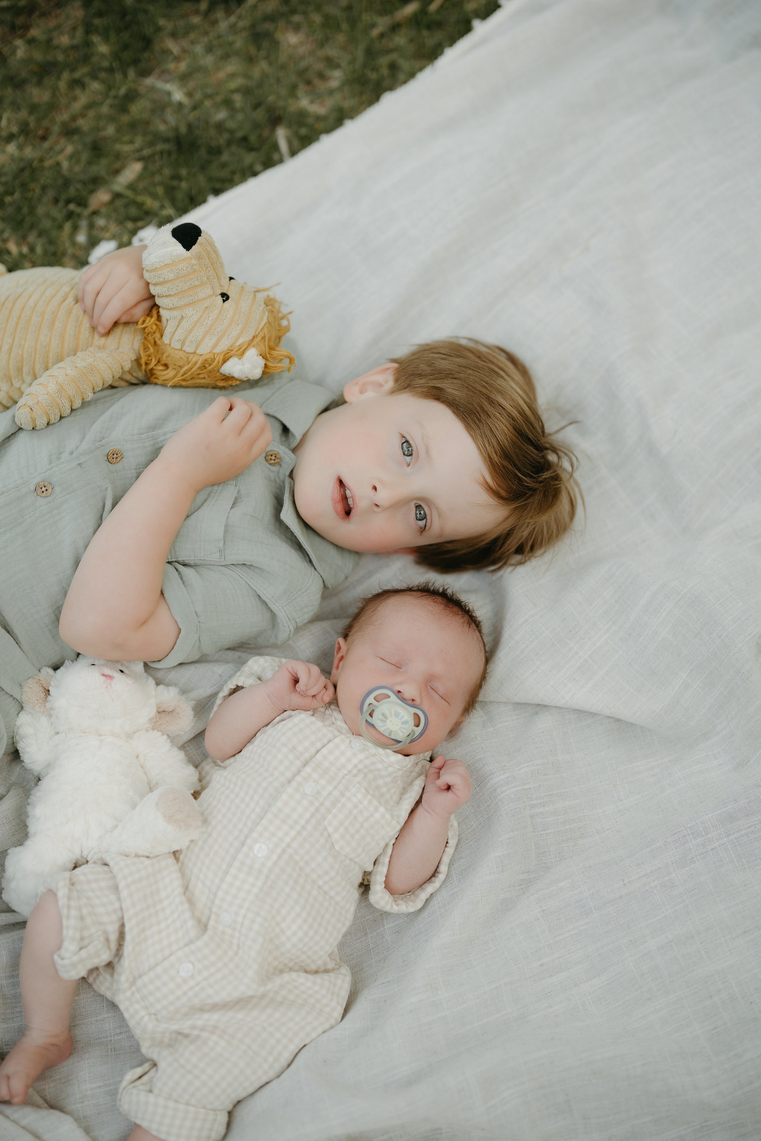 A toddler and a newborn lay on a blanket beside each other as they look up towards the camera. Calgary River Lifestyle/Family/Newborn Session with KC Photography