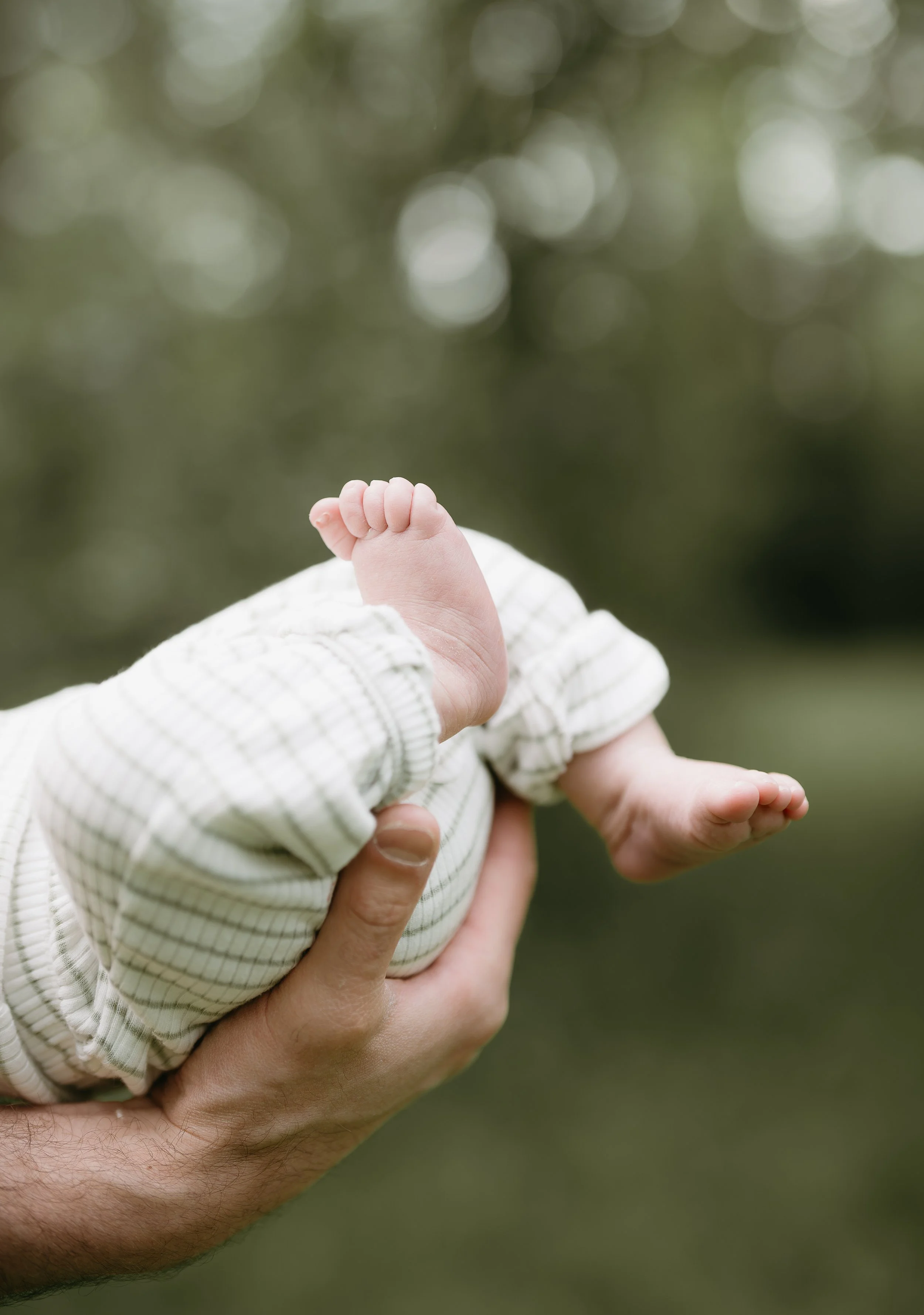 A man's hands hold the bottom of a baby who kicks their feet into the air. Calgary River Lifestyle/Family/Newborn Session with KC Photography