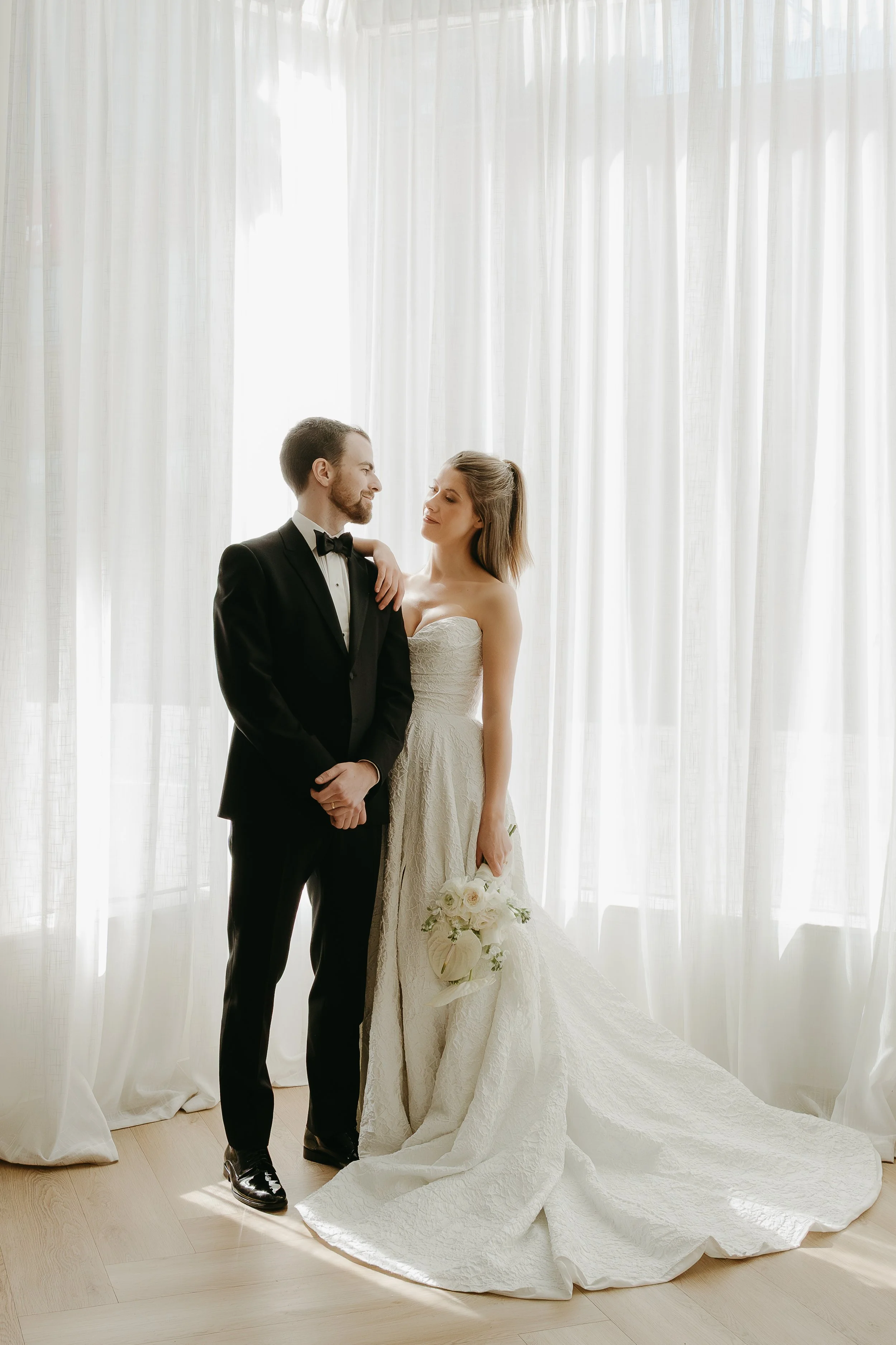 A bride and groom stand looking at one another as they are surrounded by long white curtains that drape around them. Calgary Elopement/Wedding Editorial Session with KC Photography