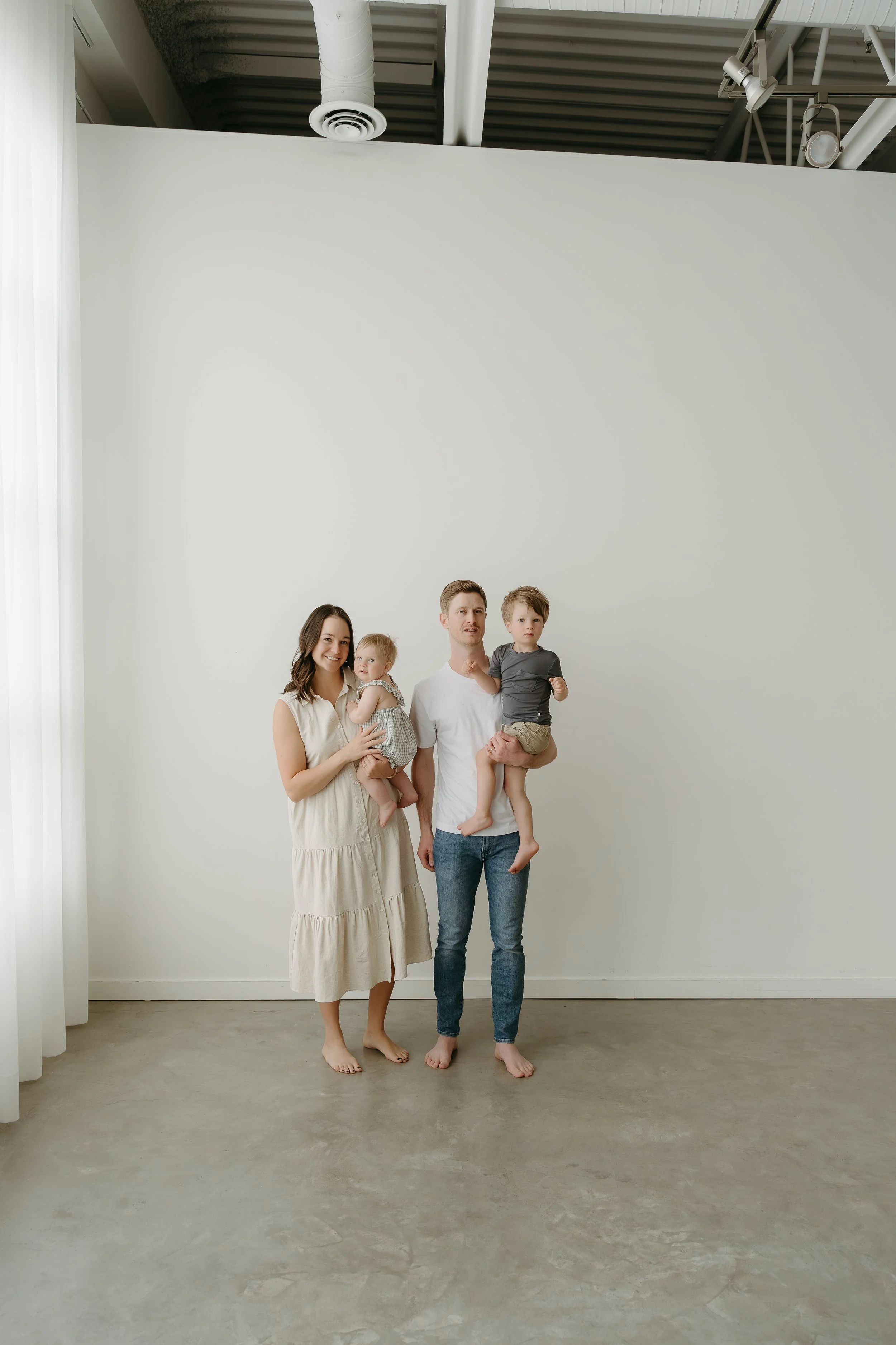 A mother and father hold their children in a studio as they stand beside each other and all look towards the camera. Calgary Lifestyle/Family Studio Session with KC Photography
