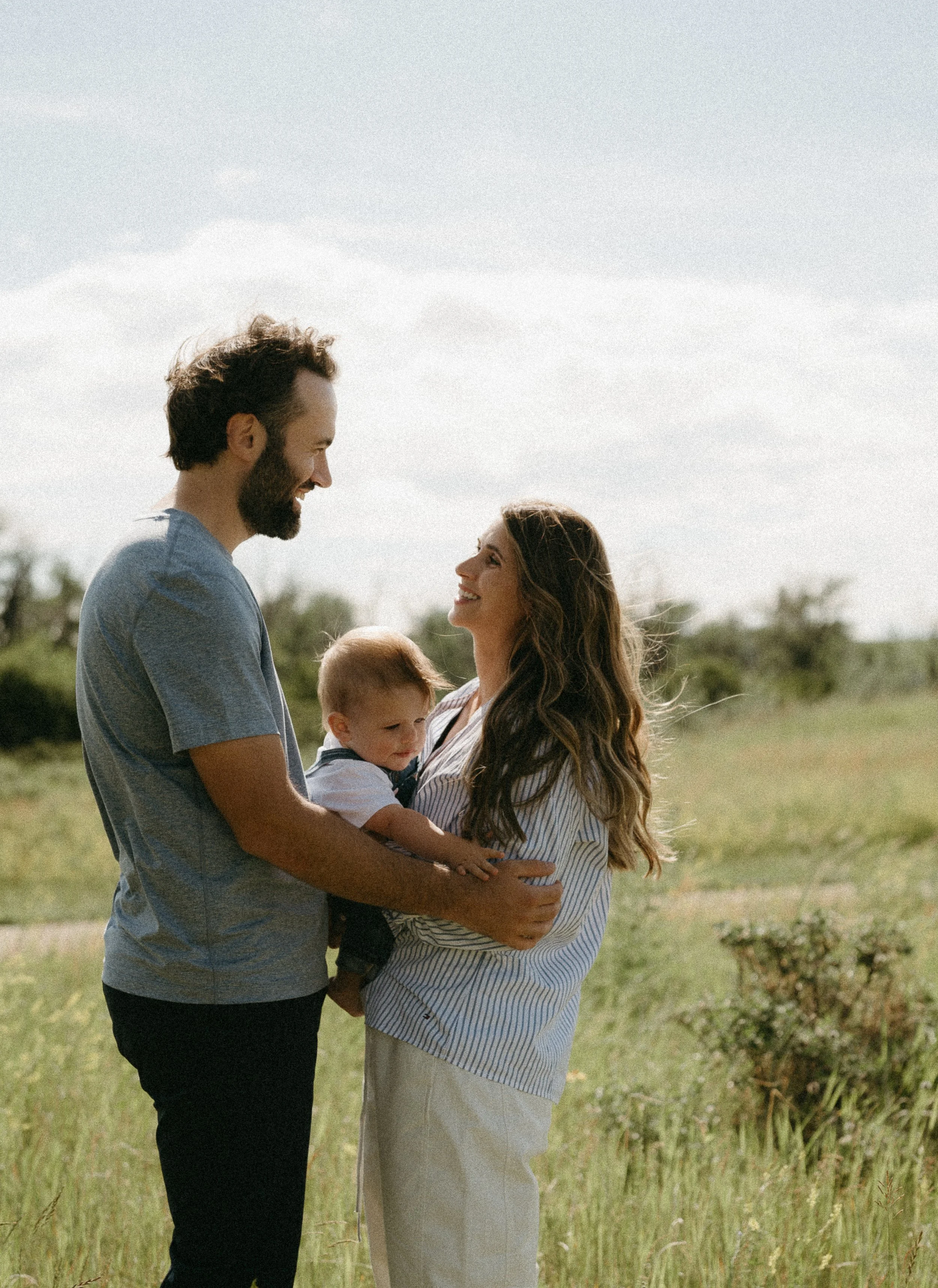A woman and man face another holding their infant together as they look at each other and smile in a field. July 2025 Calgary Lifestyle/Family Session with KC Photography