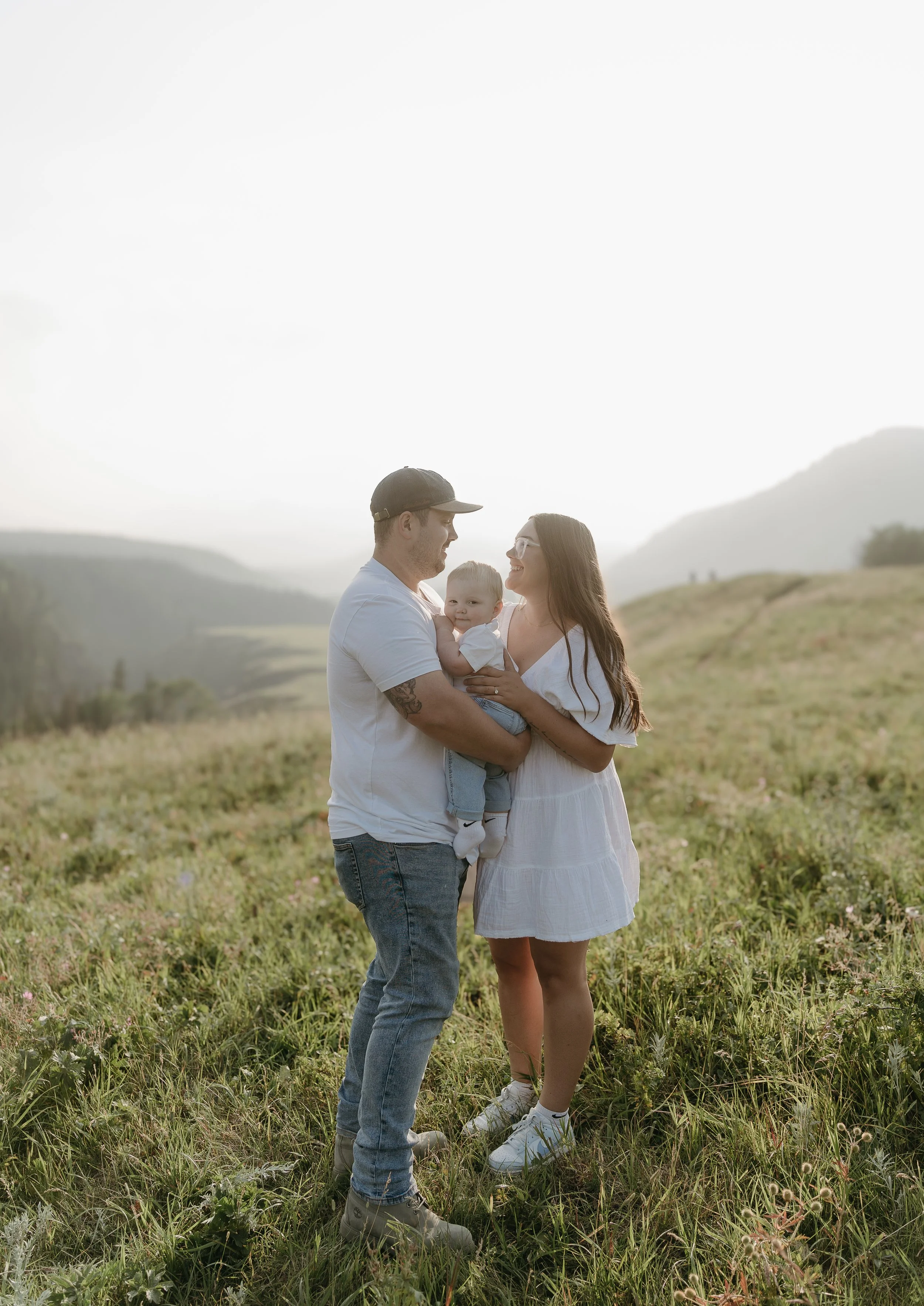 A couple stands facing each other in a field surrounded by mountains, holding their infant between them while smiling at each other. Kananaskis Lifestyle/Family Session with KC Photography