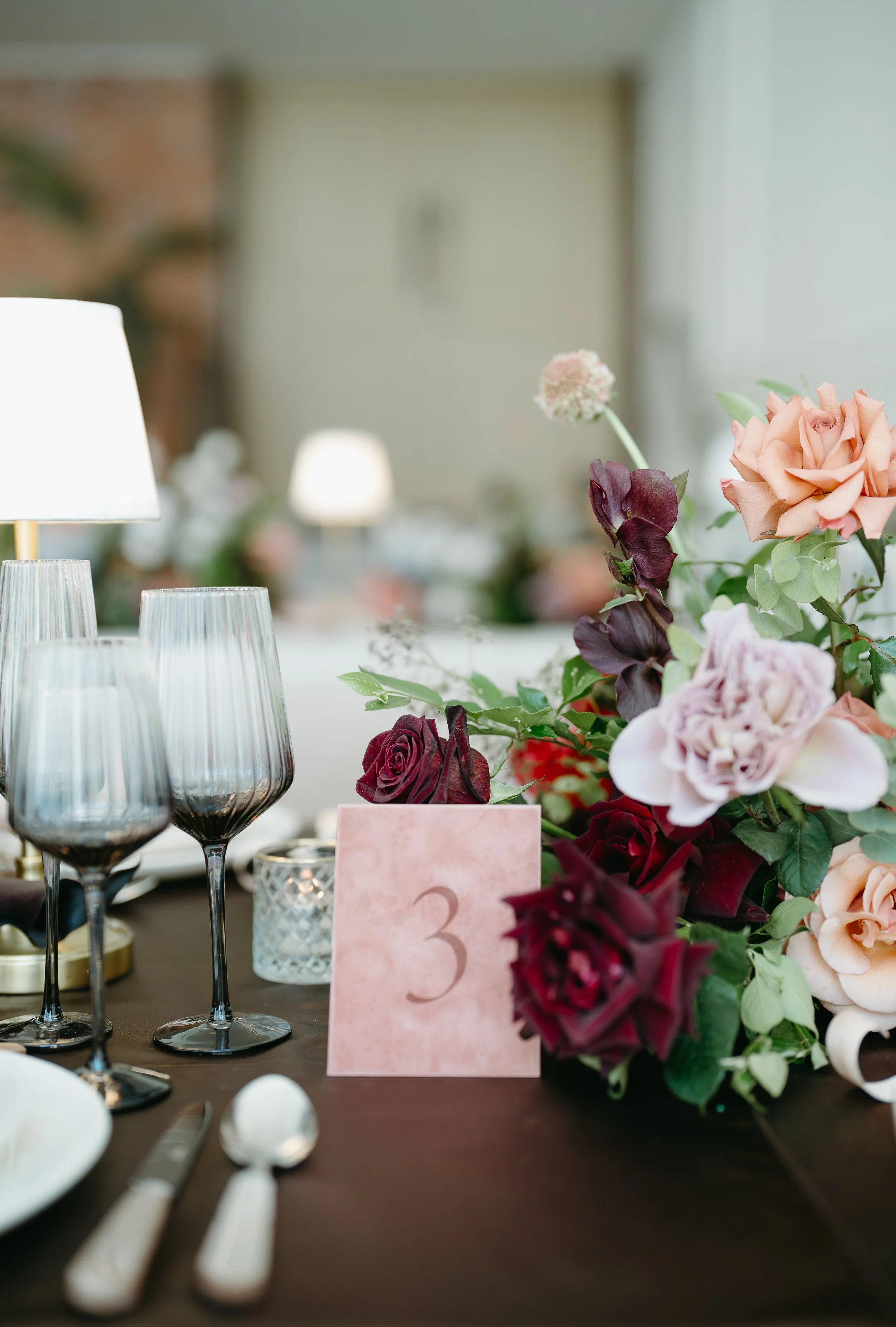 A tabletop at an empty wedding reception covered with table arrangements, bouquets of flowers, lamps, and candles. July 2025 Calgary Elopement/Wedding Editorial Session with KC Photography