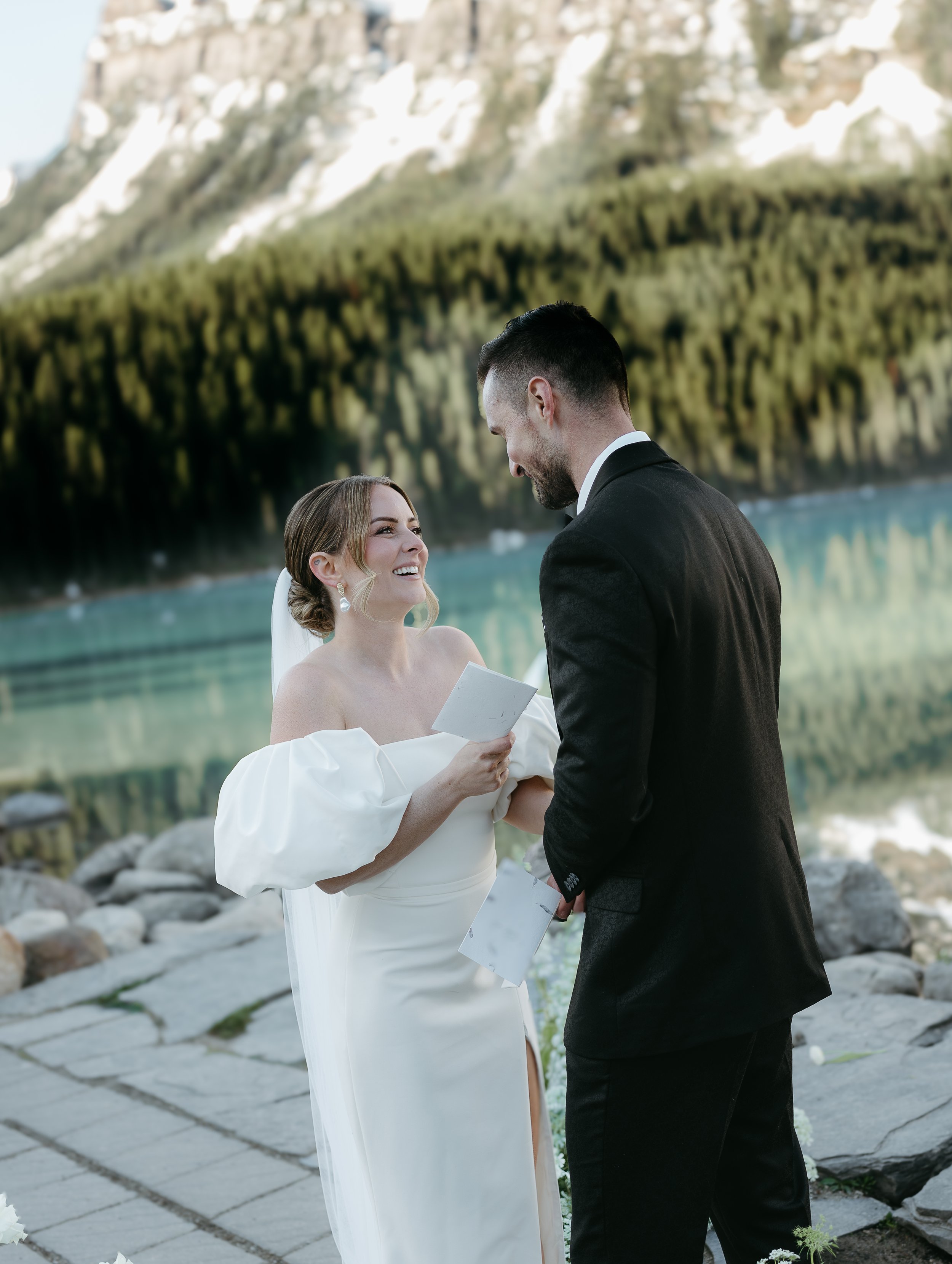 A bride and groom exchange vows on the edge of a lake surrounded by mountains. August 2025 Canmore/Banff Elopement/Wedding Editorial Session with KC Photography