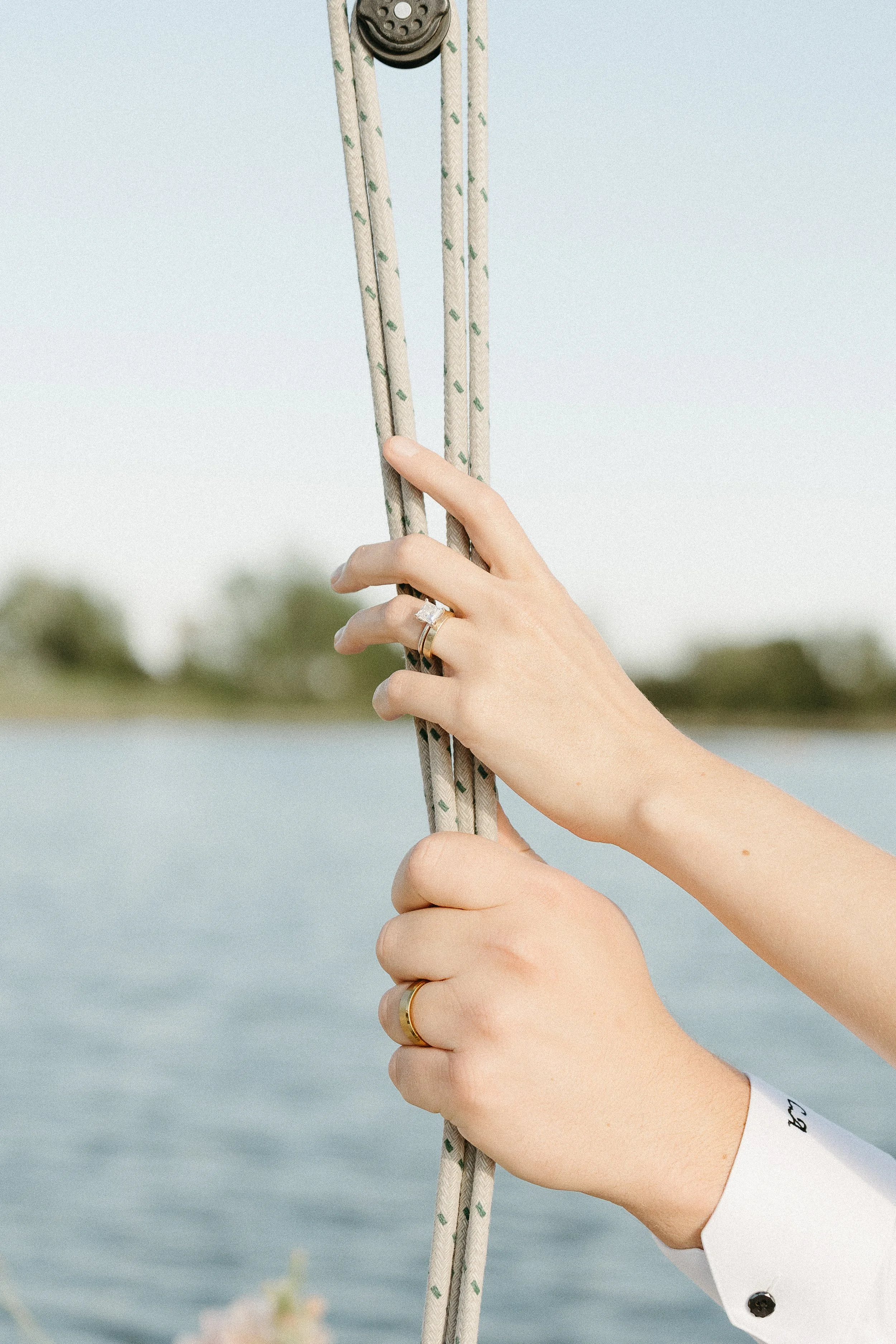 A bride and grooms hands with their wedding bands are pictured holding onto the ropes of a sailing boat. August 2025 Elopement/Wedding Sailboat/Sailing Session with KC Photography