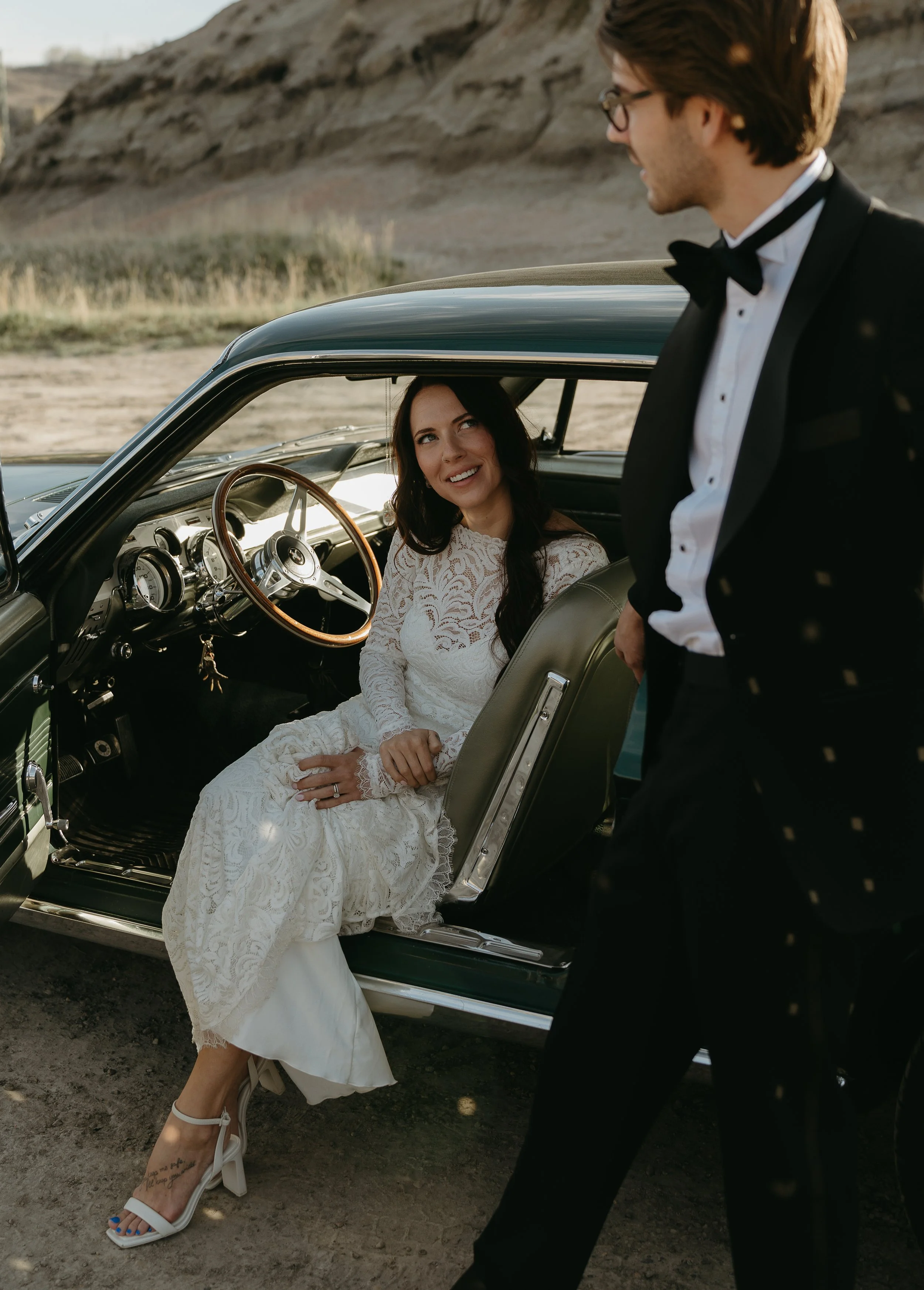 A bride sits in the passenger seat of a vintage car, gazing up at her husband standing beside the car in Drumheller. April 2025 Drumheller Elopement/Wedding Session with KC Photography