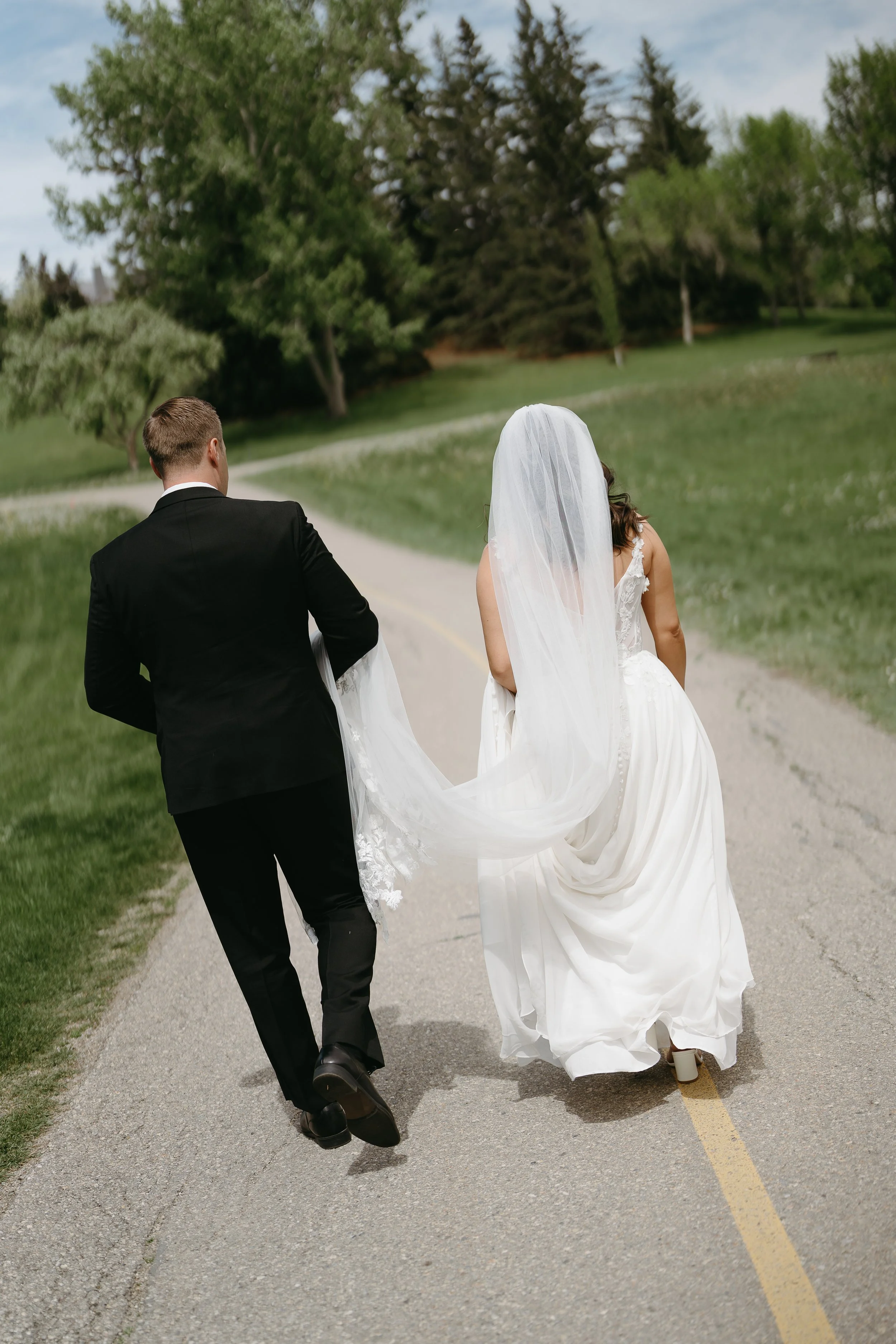 A bride and groom walk down a pathway in a park while the bride holds her wedding gown and the groom holds the bottom of her veil. Jessica and Devon May 2025 Wedding Photography Session with KC Photography