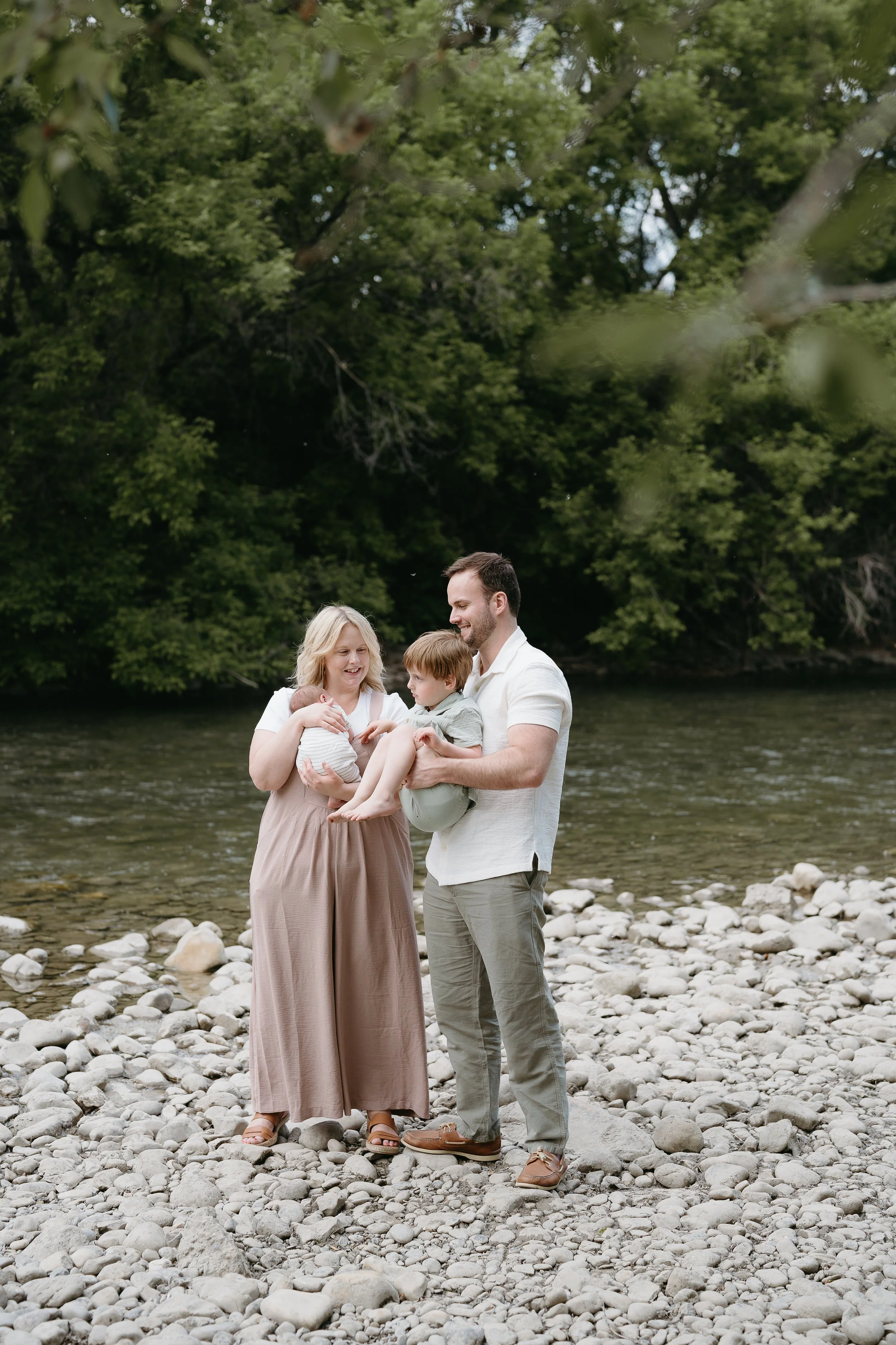 A man and woman hold each of their children as they stand along the side of a riverbank. Calgary River Lifestyle/Family/Newborn Session with KC Photography