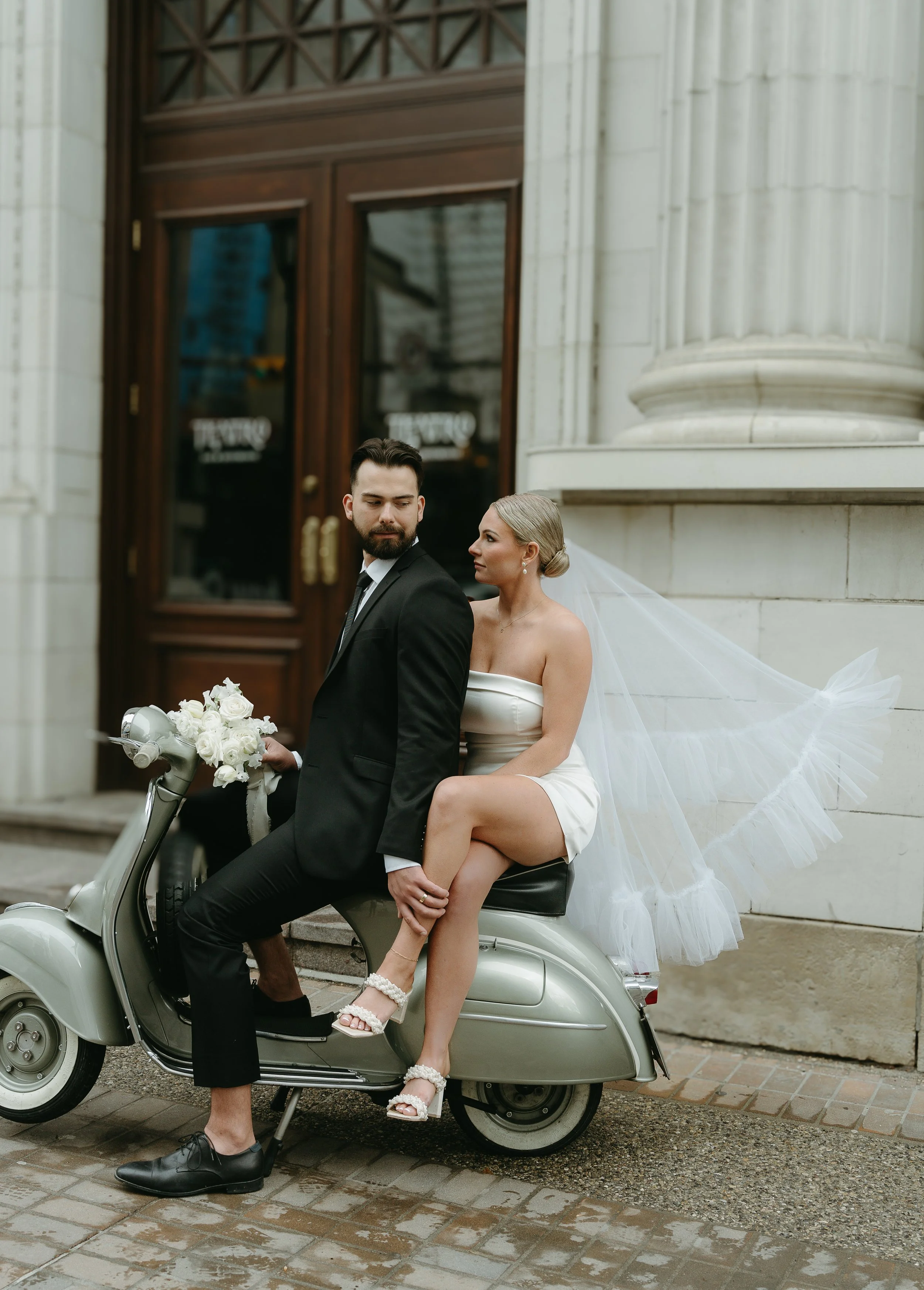 A bride and groom sit on a Moped motorcycle while the bride's wedding veil flows in the wind out front of Teatro in Downtown Calgary. Downtown Calgary Elopement/Wedding Session with KC Photography