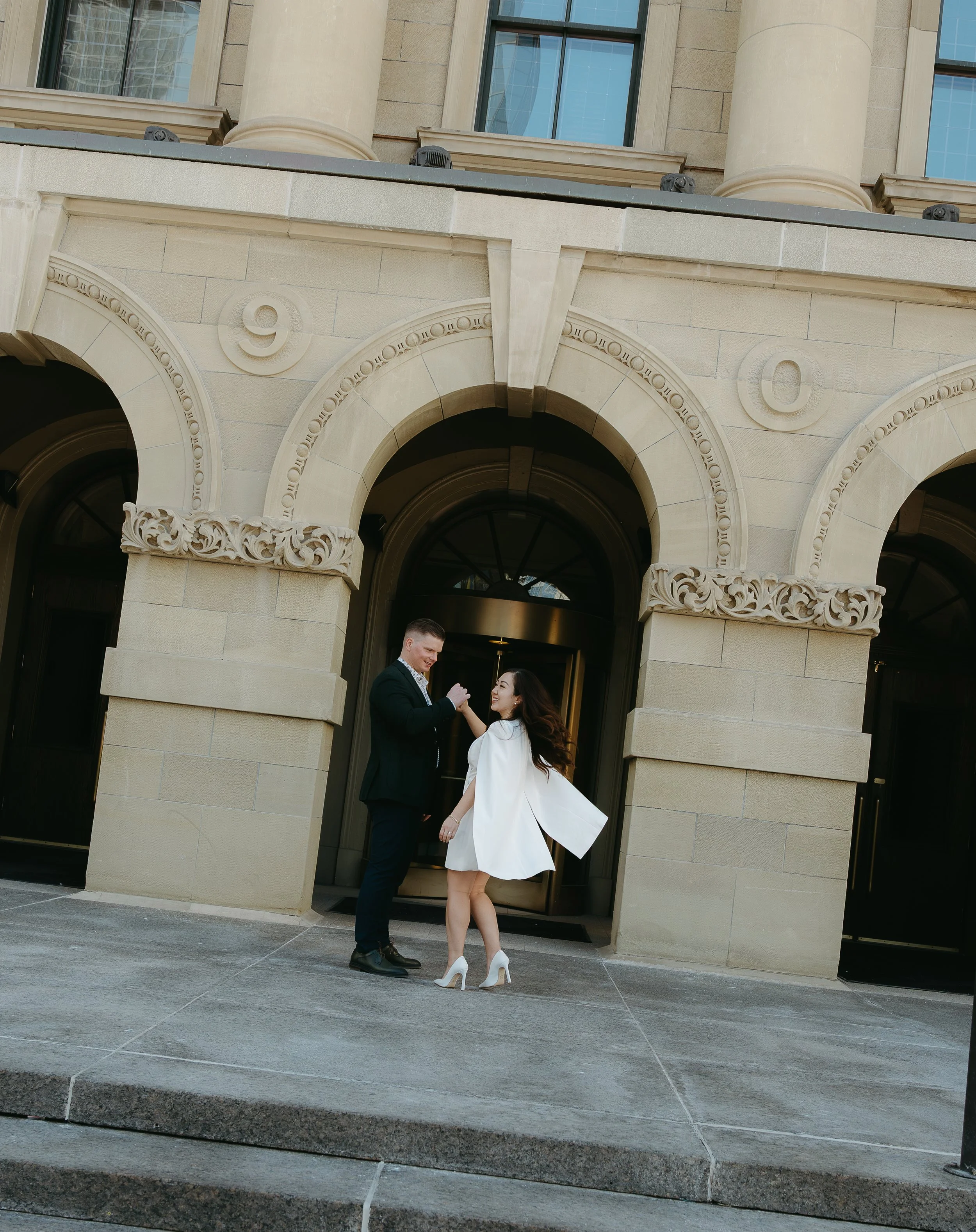 A man twirls his fiancé on the top of stairs outside a building in Downtown Calgary. Downtown Calgary Engagement Session with KC Photography