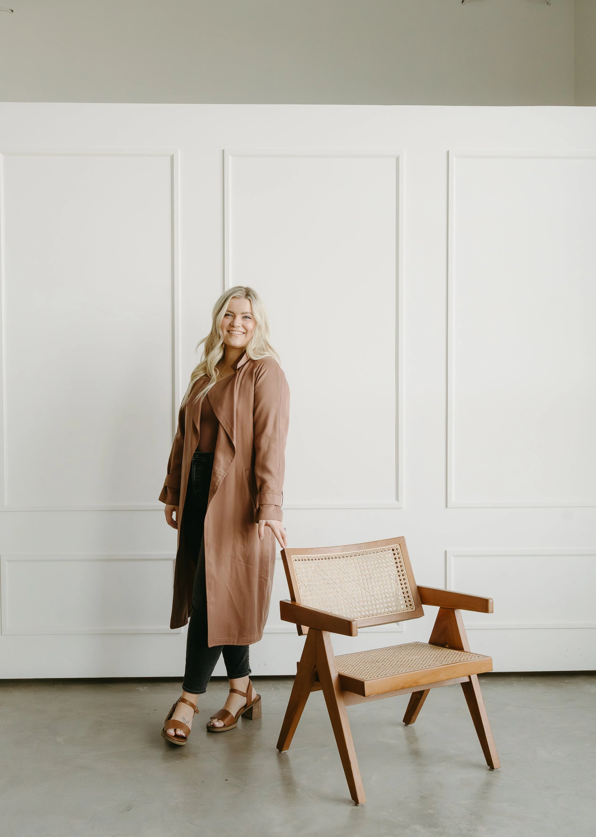 A woman approaches from behind a wooden chair while looking at the camera smiling. Calgary Studio Branding Session with KC Photography
