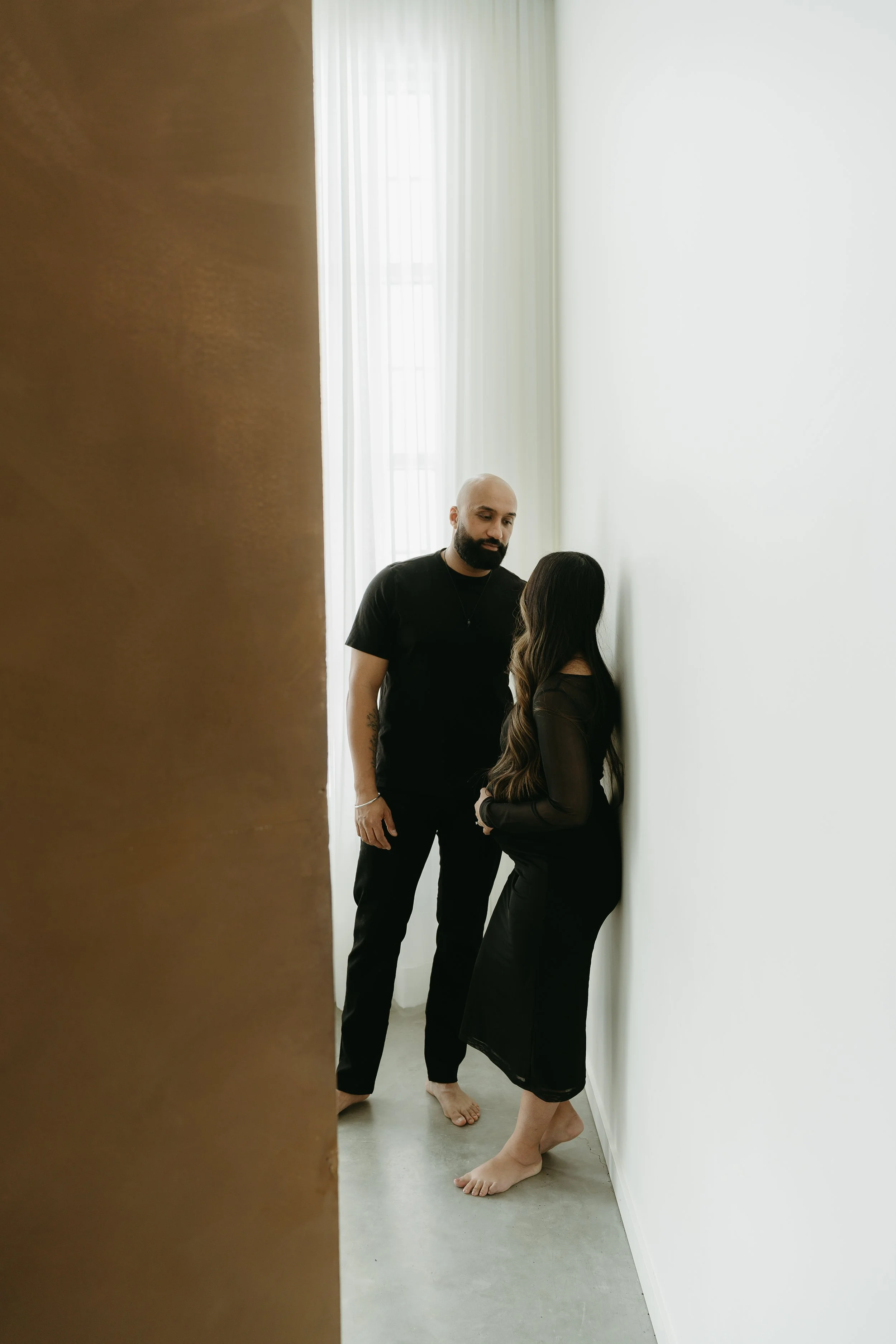 A man looks at his pregnant wife who is pregnant and holding her belly, while leaning on the white wall of a studio. Calgary Lifestyle/Family/Newborn/Pregnancy/Maternity Studio Session with KC Photography