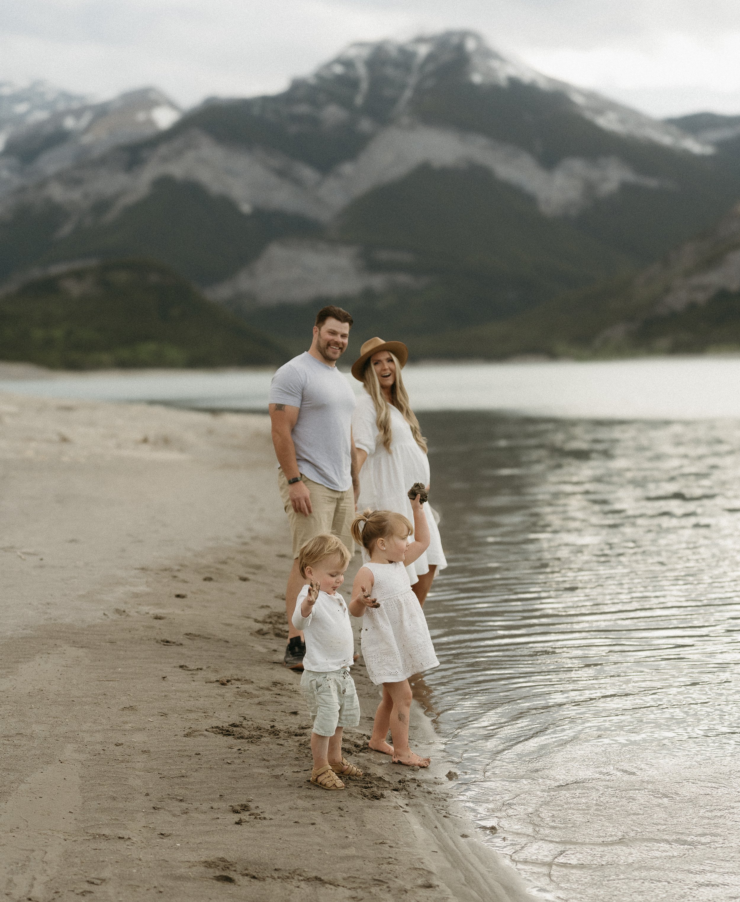 A couple stands watching their two toddles play in the sand along the bank of a river surrounded by mountains. Canmore/Banff Lifestyle/Family Session with KC Photography