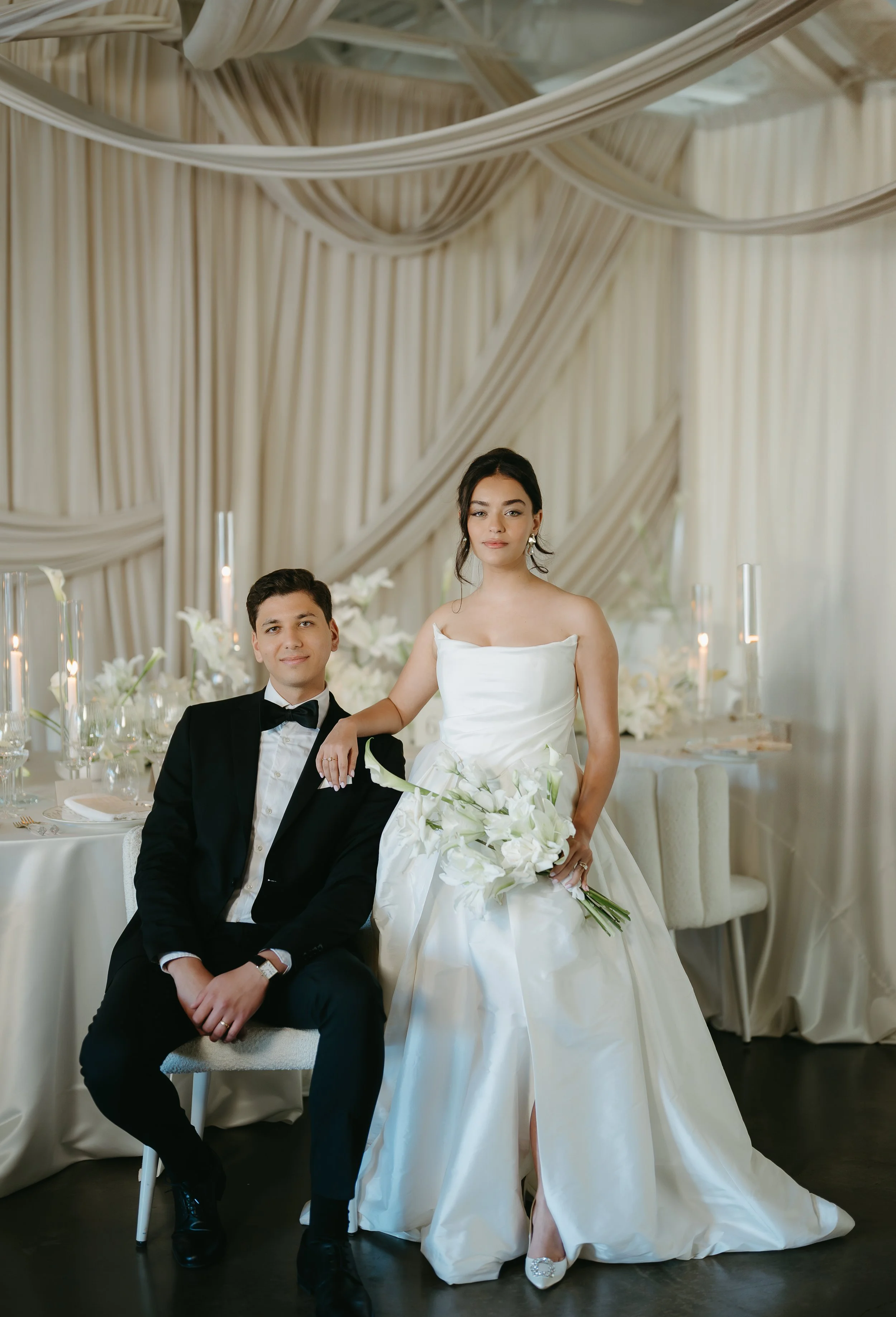 A groom sits in a chair while his bride stands beside him in their empty reception. July 2025 Calgary Elopement/Wedding Editorial Session with KC Photography