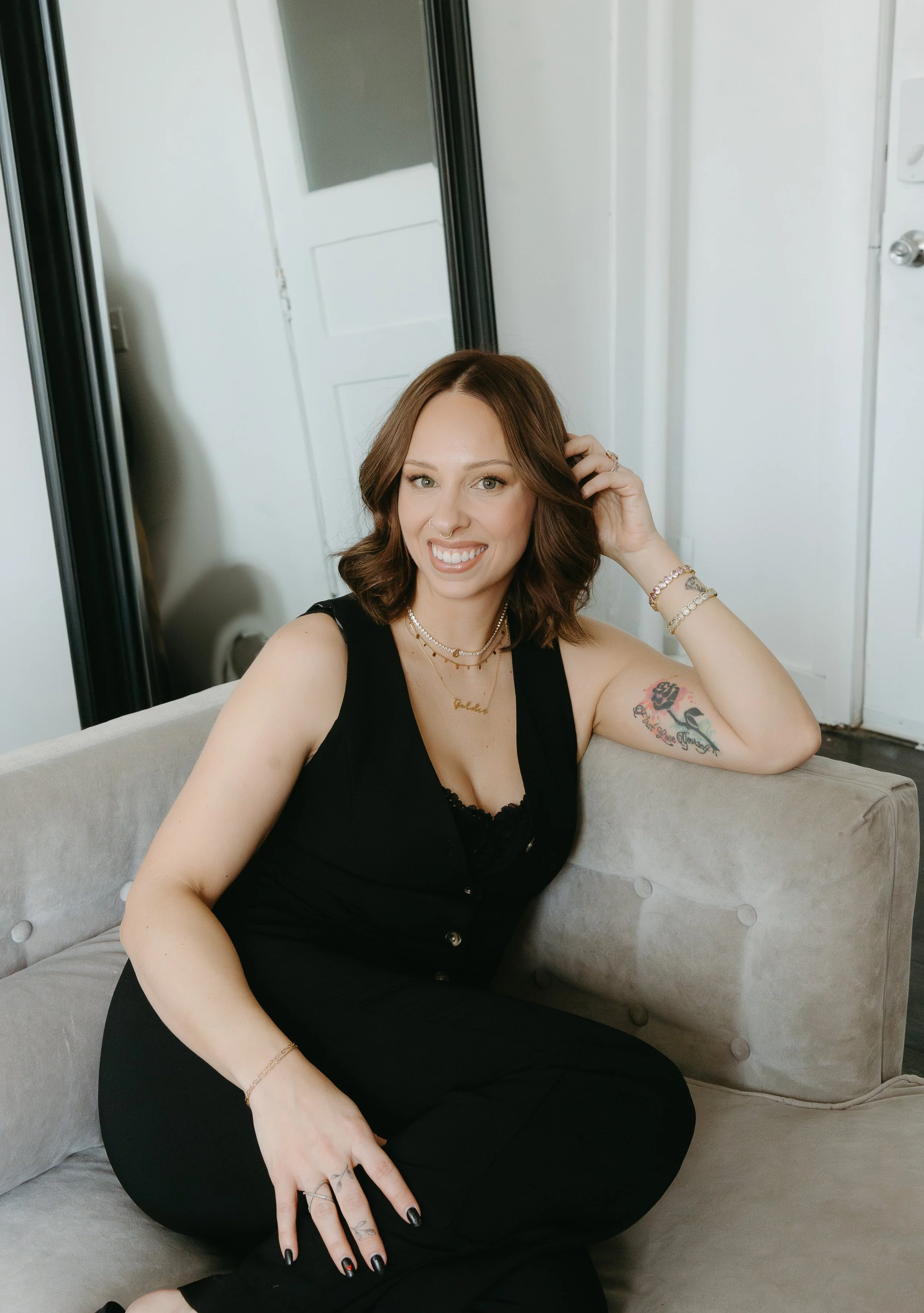 A woman sits in the corner of a couch and rests her arm on the edge while touching her hair and smiling at the camera. Beauty Ave Studio Branding Session with KC Photography