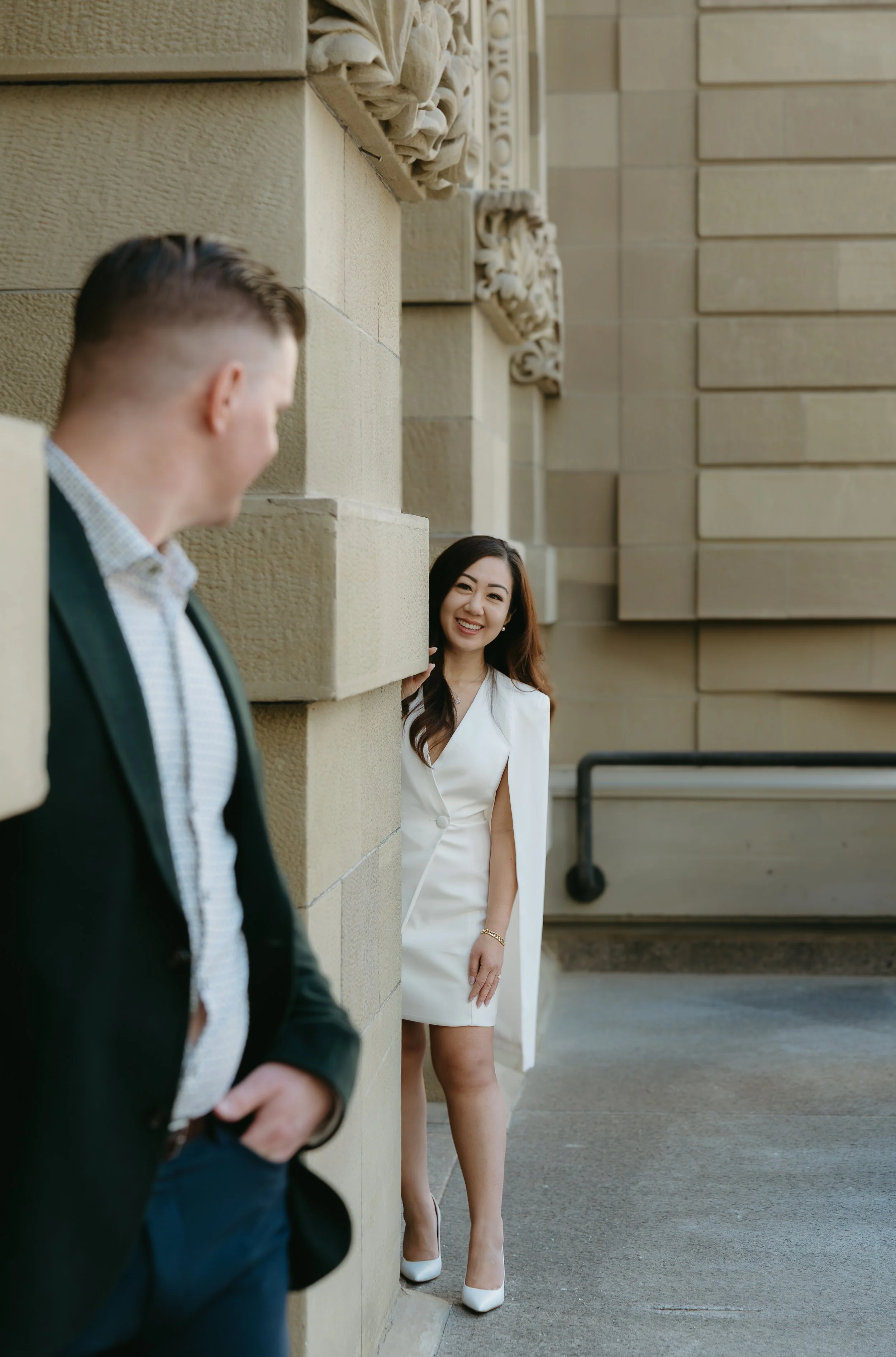A fiancé appears from a row of pillars walking towards her fiancé in Downtown Calgary. Downtown Calgary Engagement Session with KC Photography