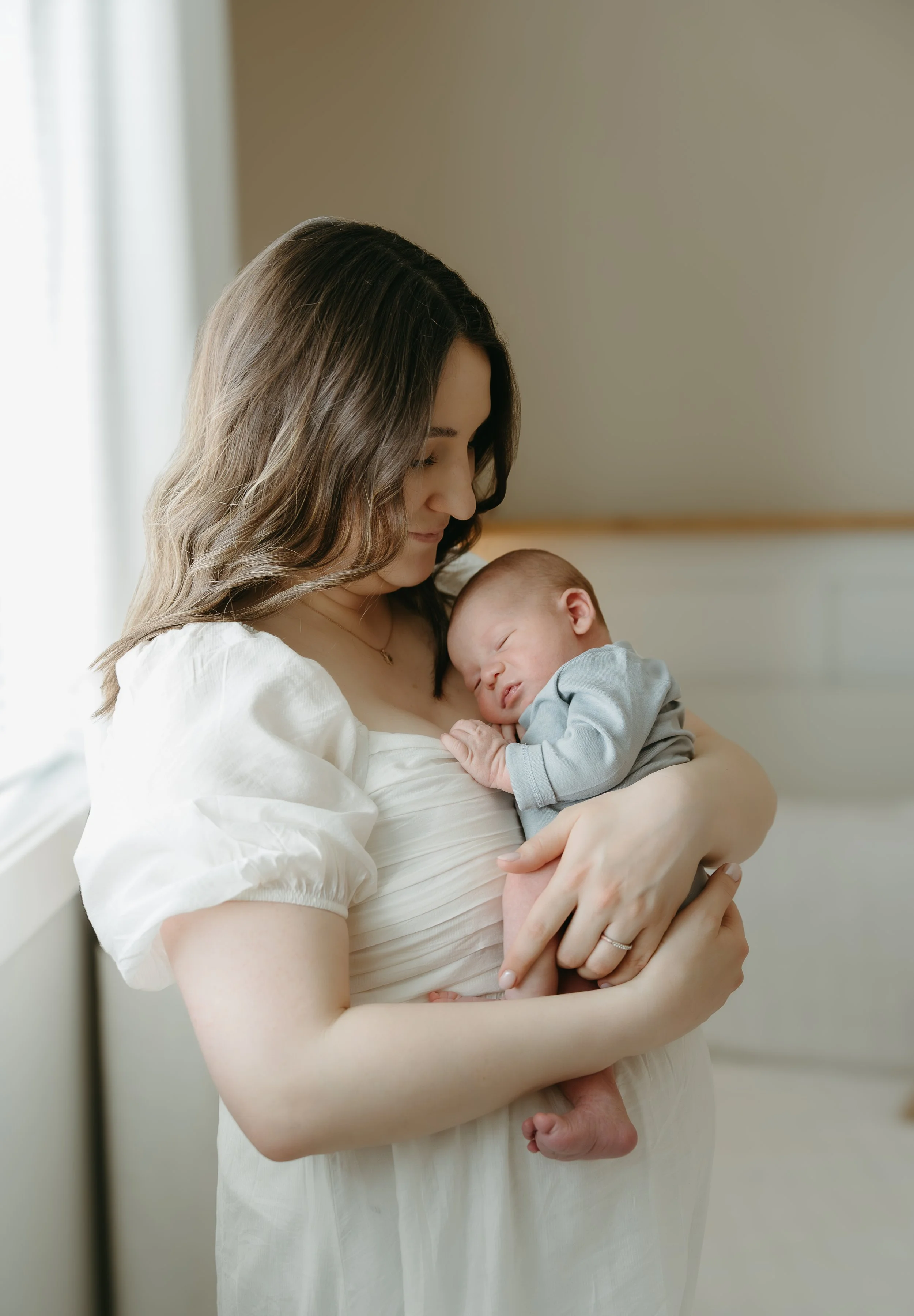 A woman looks down at her newborn baby she is holding that is cuddled into her chest. Calgary Home Lifestyle/Family/Newborn Session with KC Photography