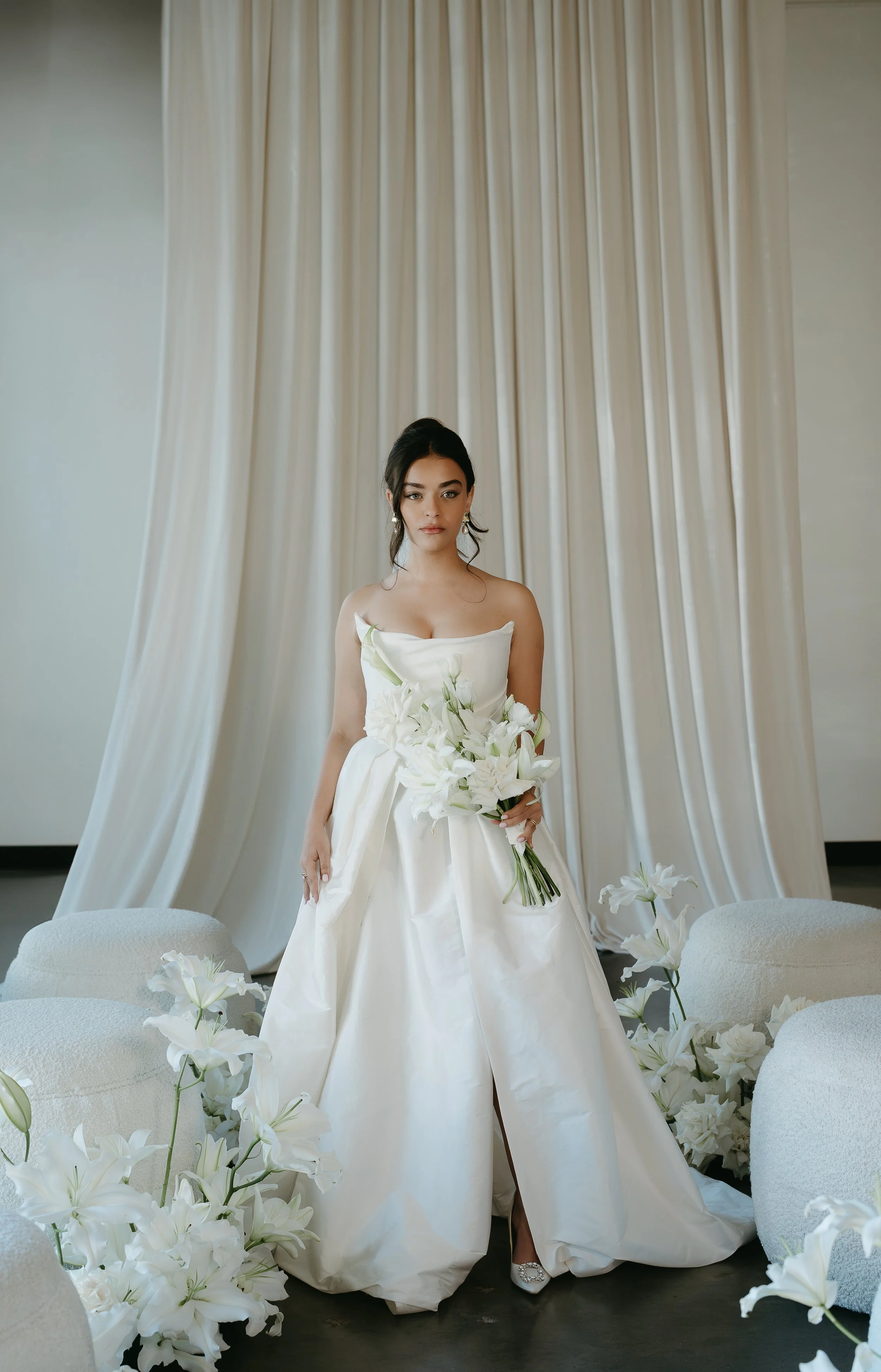 A bride stands in an aisle of chairs and white lily's holding a bouquet. July 2025 Calgary Elopement/Wedding Editorial Session with KC Photography