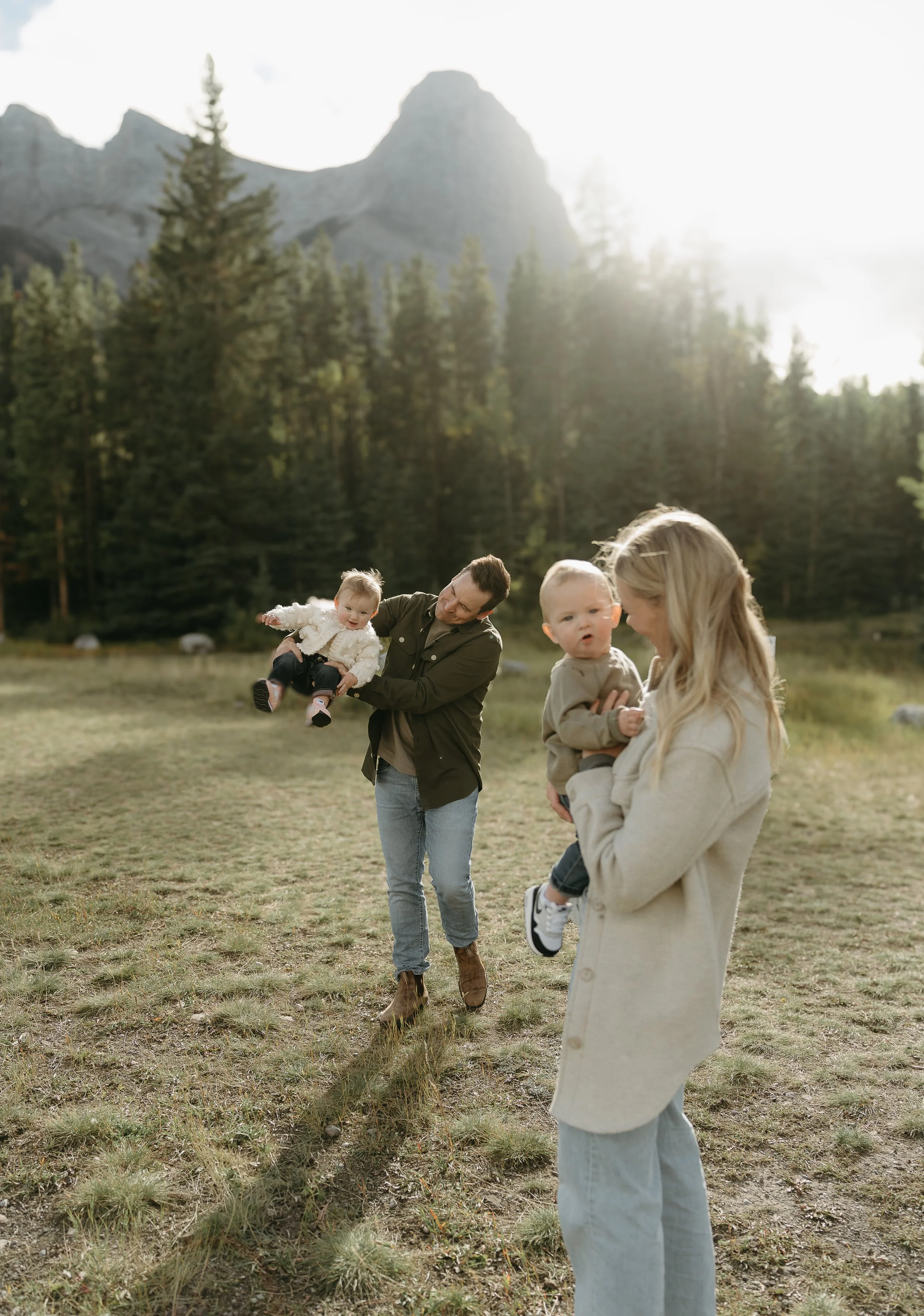 A women holds her infant and looks at them smiling while her husband playfully swings their other infant around in the background of a field surrounded by mountains. Kananaskis Lifestyle/Family Session with KC Photography