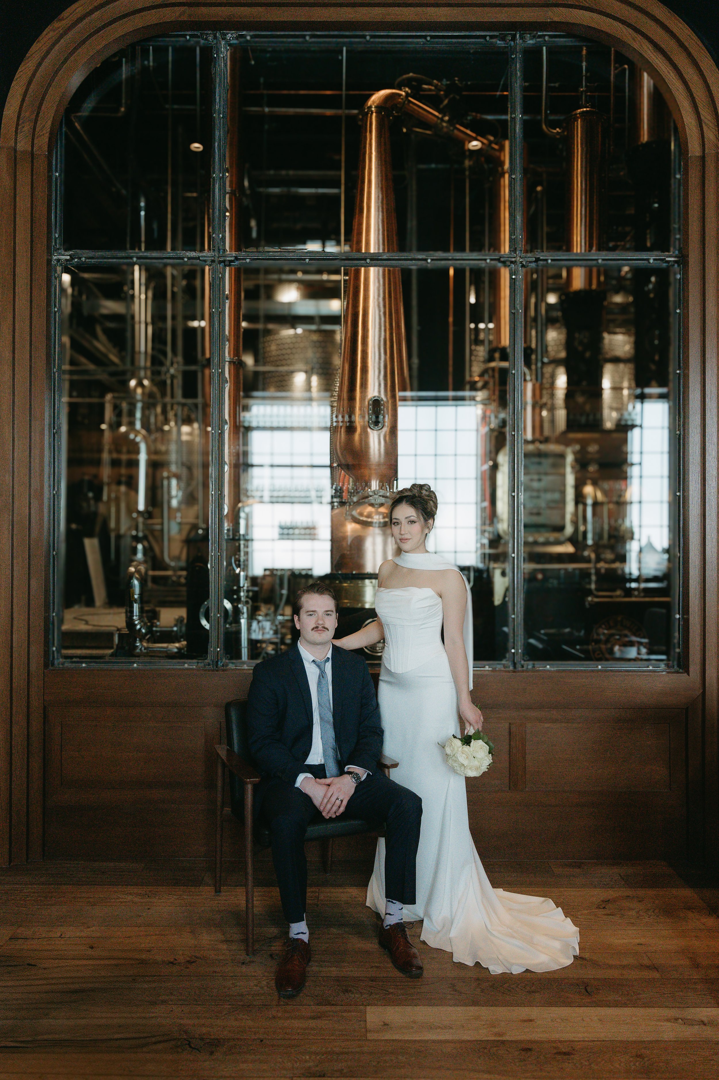 A bride stands closely beside her seated groom while they both gaze into the camera. Calgary Elopement/Wedding Session with KC Photography