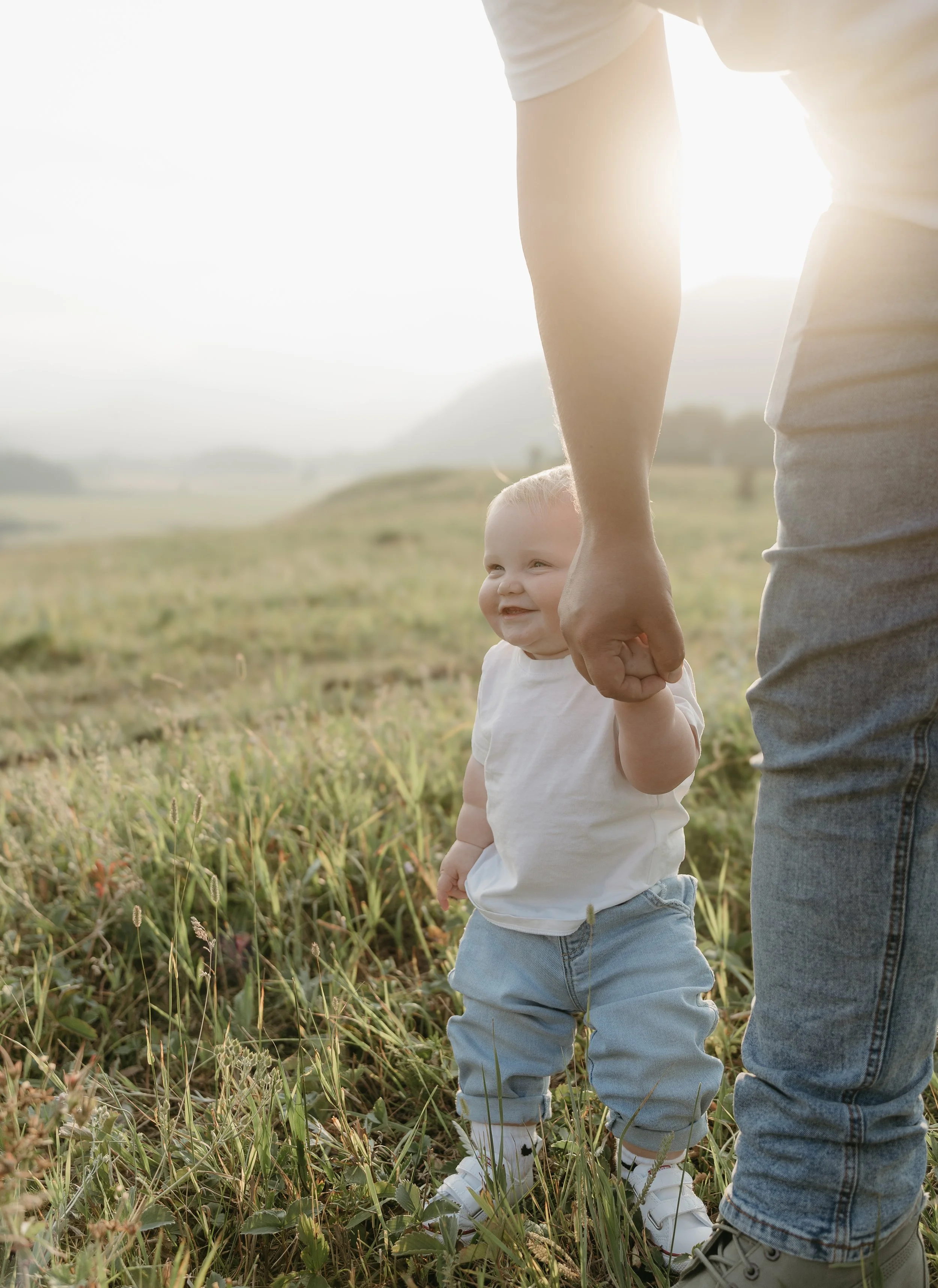 An infant holds his fathers hand as he smiles, walking through a field surrounded by mountains. Kananaskis Lifestyle/Family Session with KC Photography