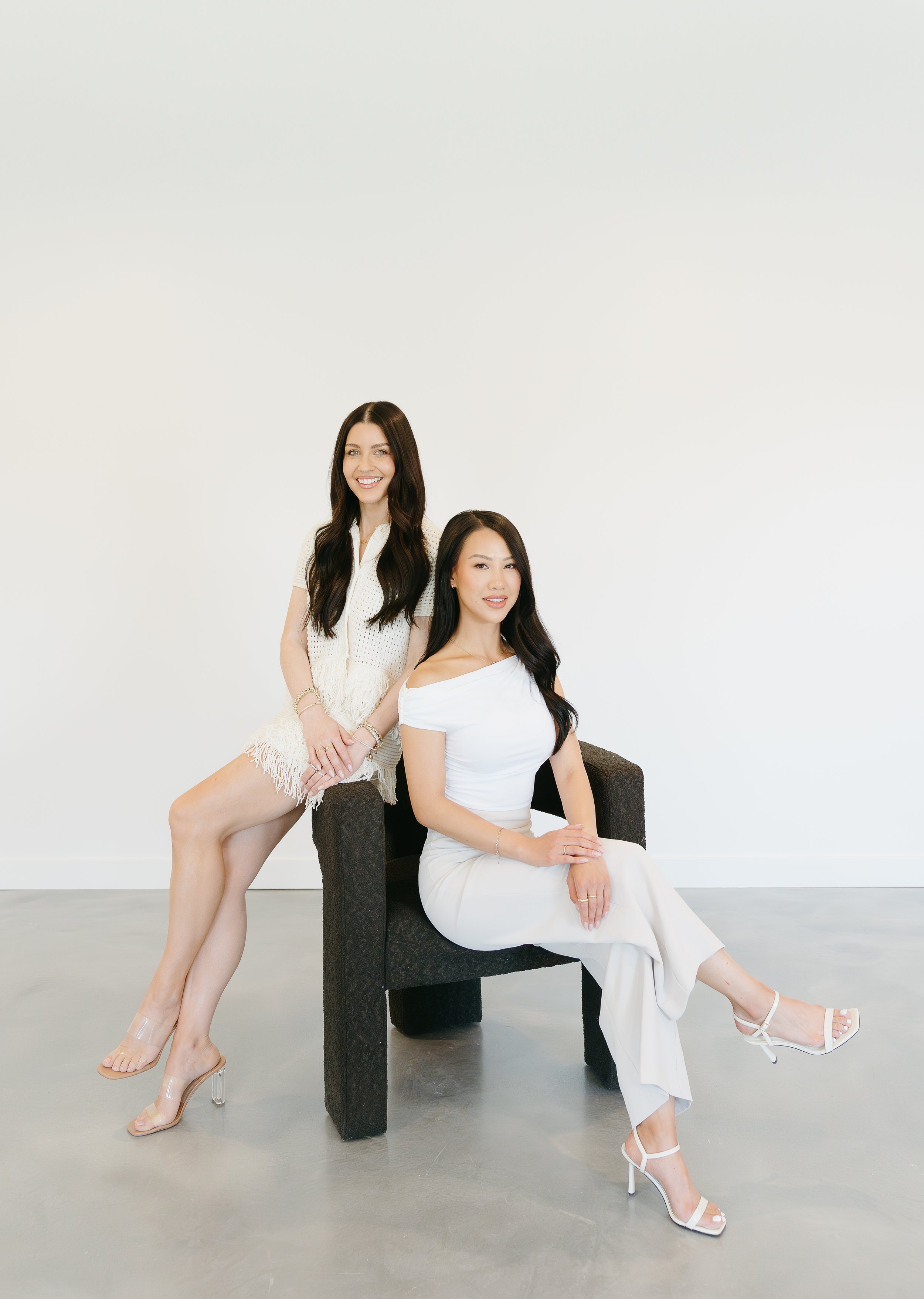 A woman is sat on a chair while another woman sits on the arm of the chair, both of them crossing their legs out and looking into the camera smiling. Beauty Ave Studio Branding Session with KC Photography