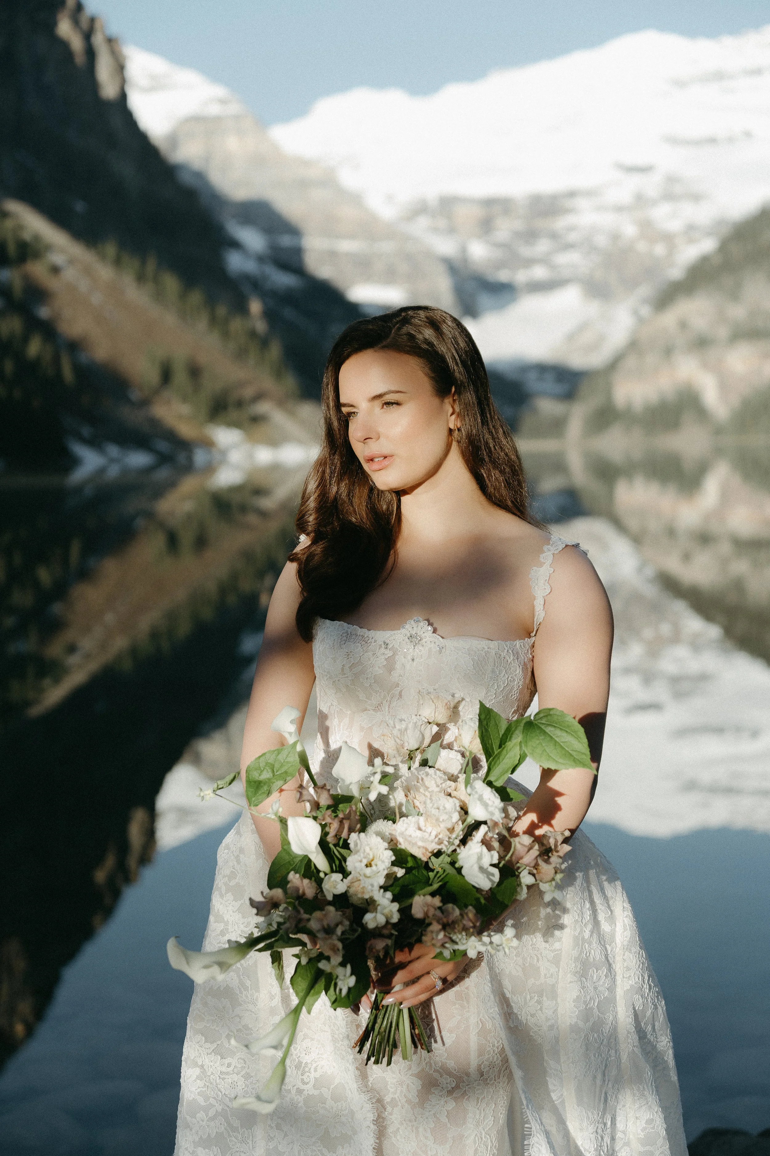 A bride holds her bouquet as Lake Louise and mountains are pictured in the background.June 2025 Lake Louise Elopement/Wedding Editorial Session with KC Photography