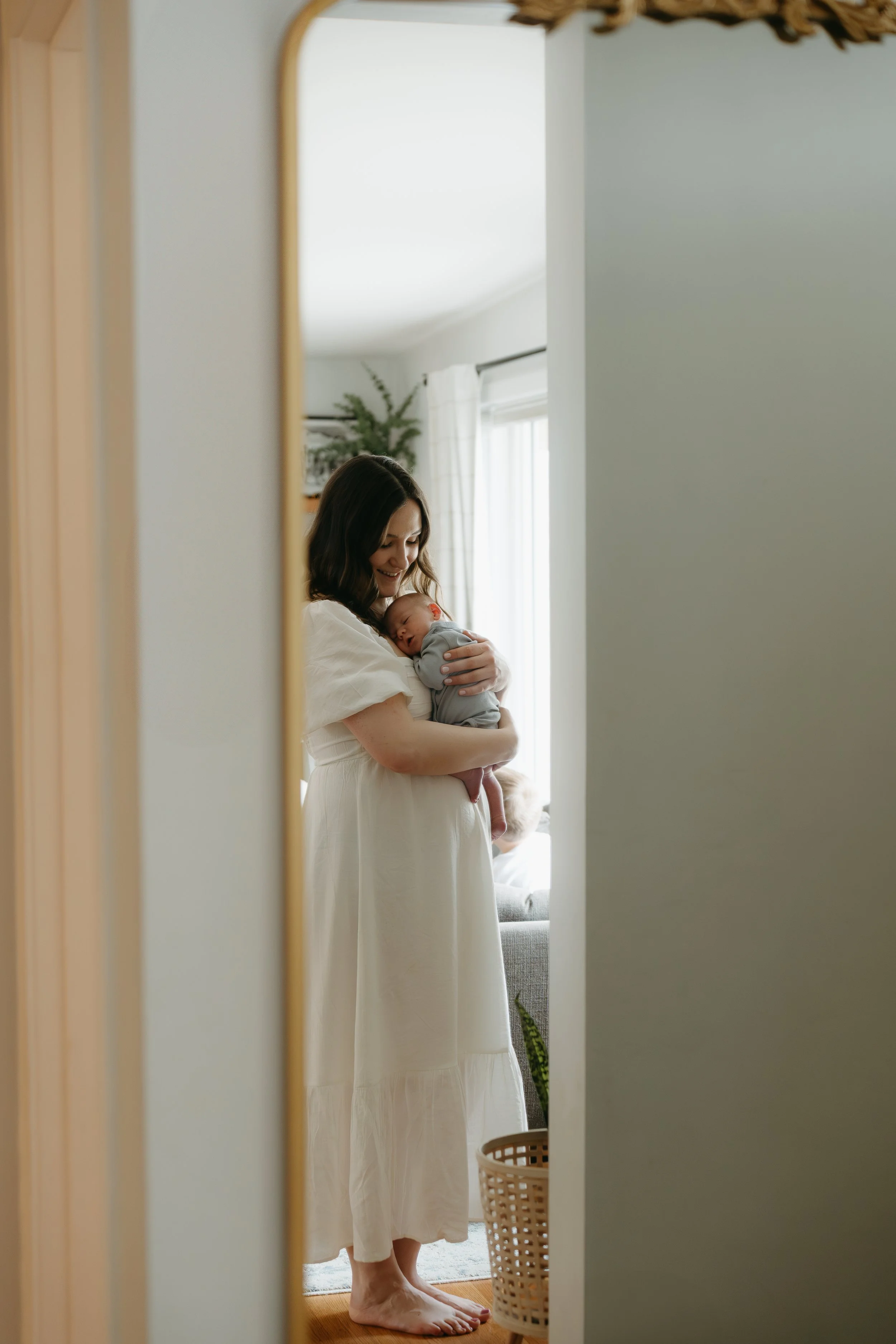 A woman is captured through a mirror looks down at her newborn baby she is holding that is cuddled into her chest. Calgary Home Lifestyle/Family/Newborn Session with KC Photography