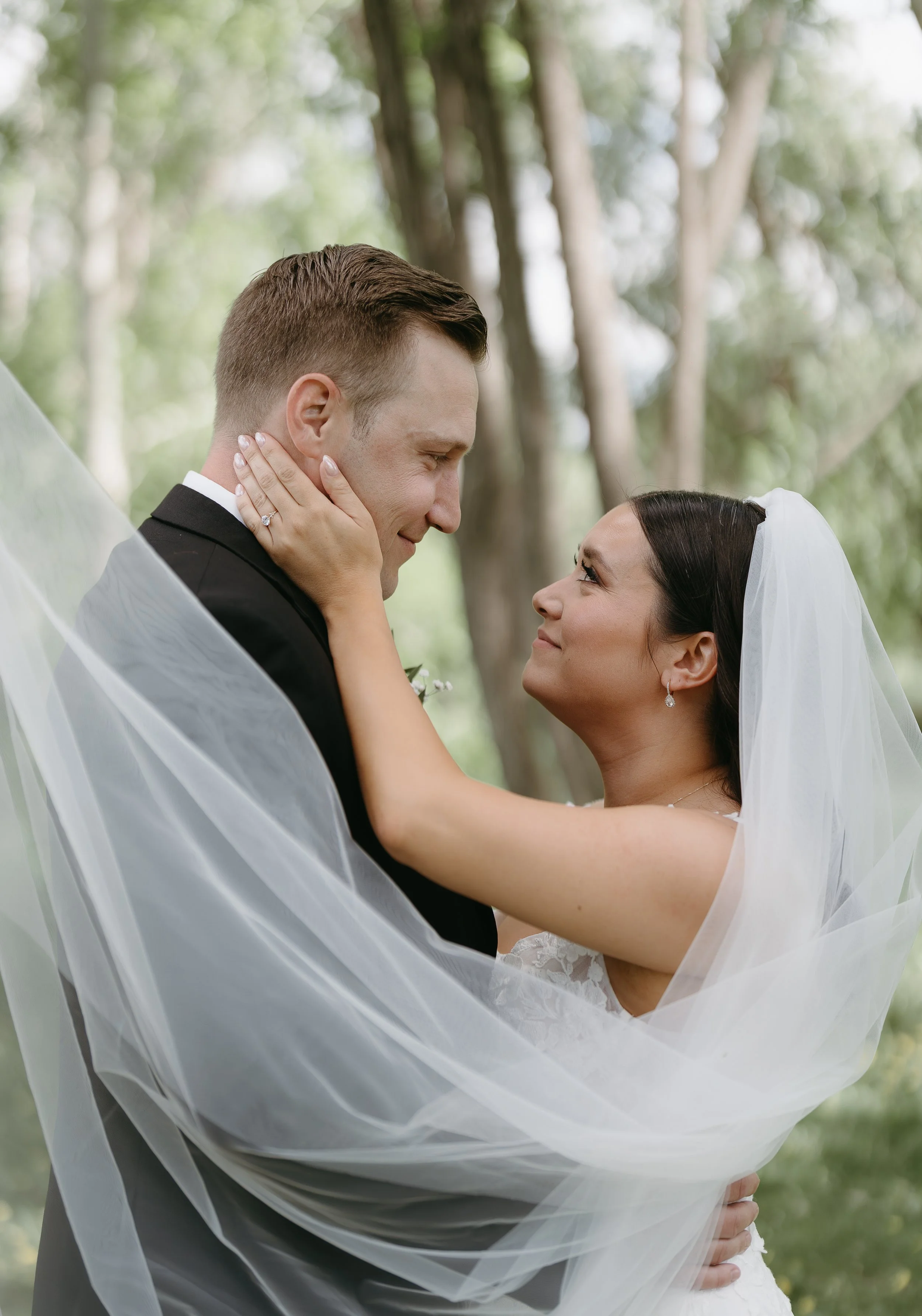 A bride places her hand on the cheek of her husband as they gaze into each others eyes in a field while her wedding veil blows in the wind. Jessica and Devon May 2025 Wedding Photography Session with KC Photography