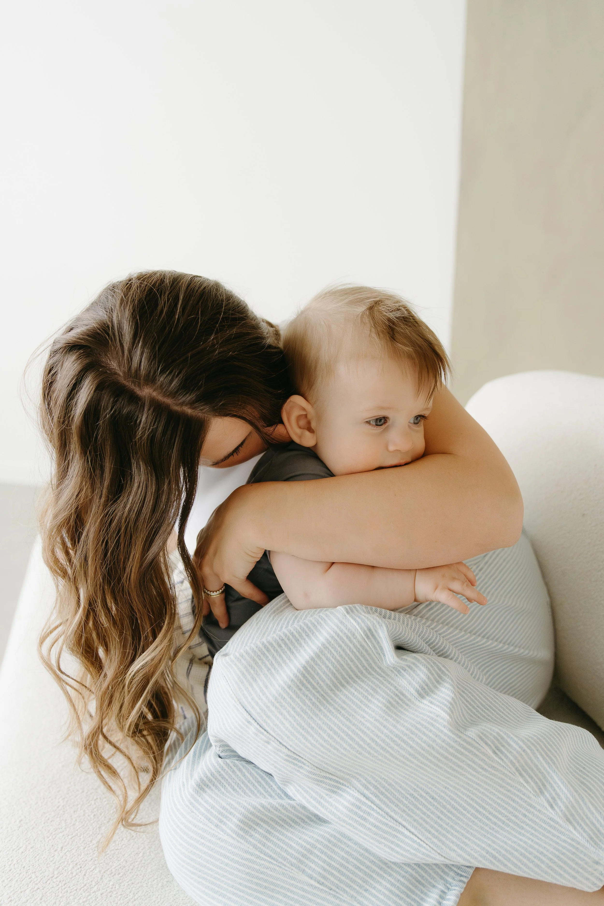 A mother wraps herself around her infant and kisses their head. Calgary Lifestyle/Infant/Family Studio Session with KC Photography