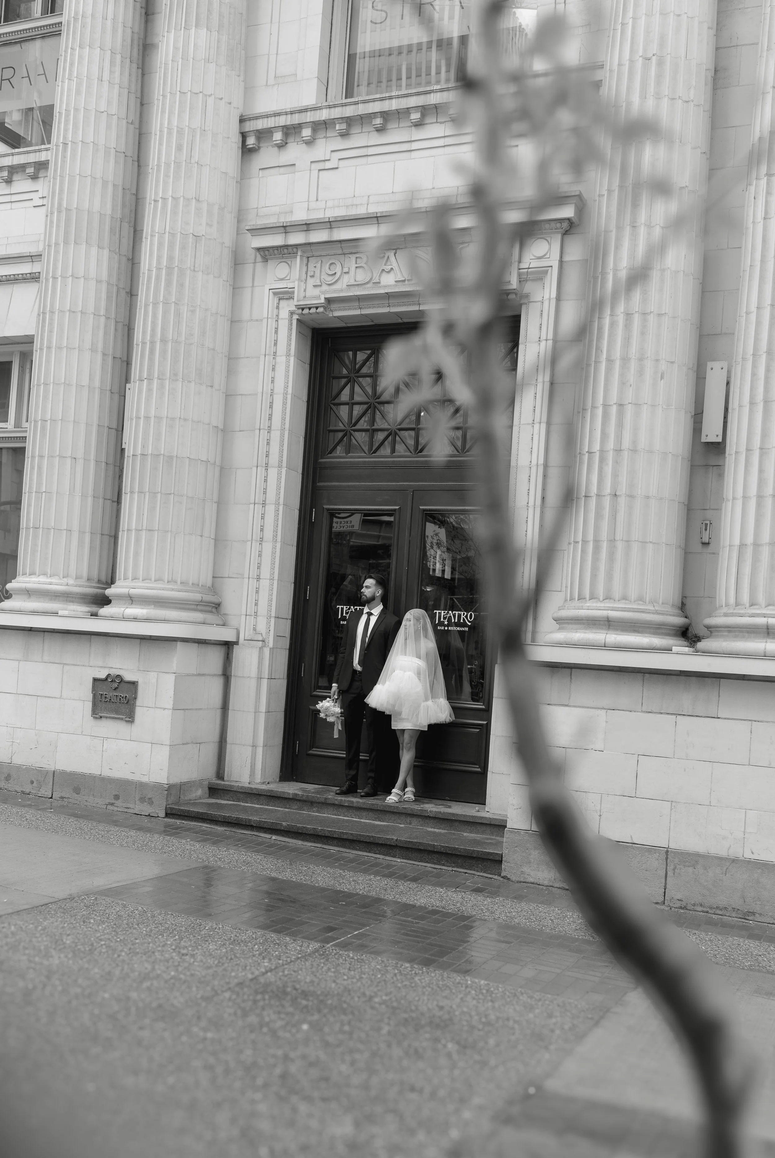A man and woman in wedding attire standing outside a building with columns and a sign that says 'TEATRO'. The man is holding a bouquet of flowers and the woman is wearing a white dress with a veil. The photo is in black and white with the text 'where