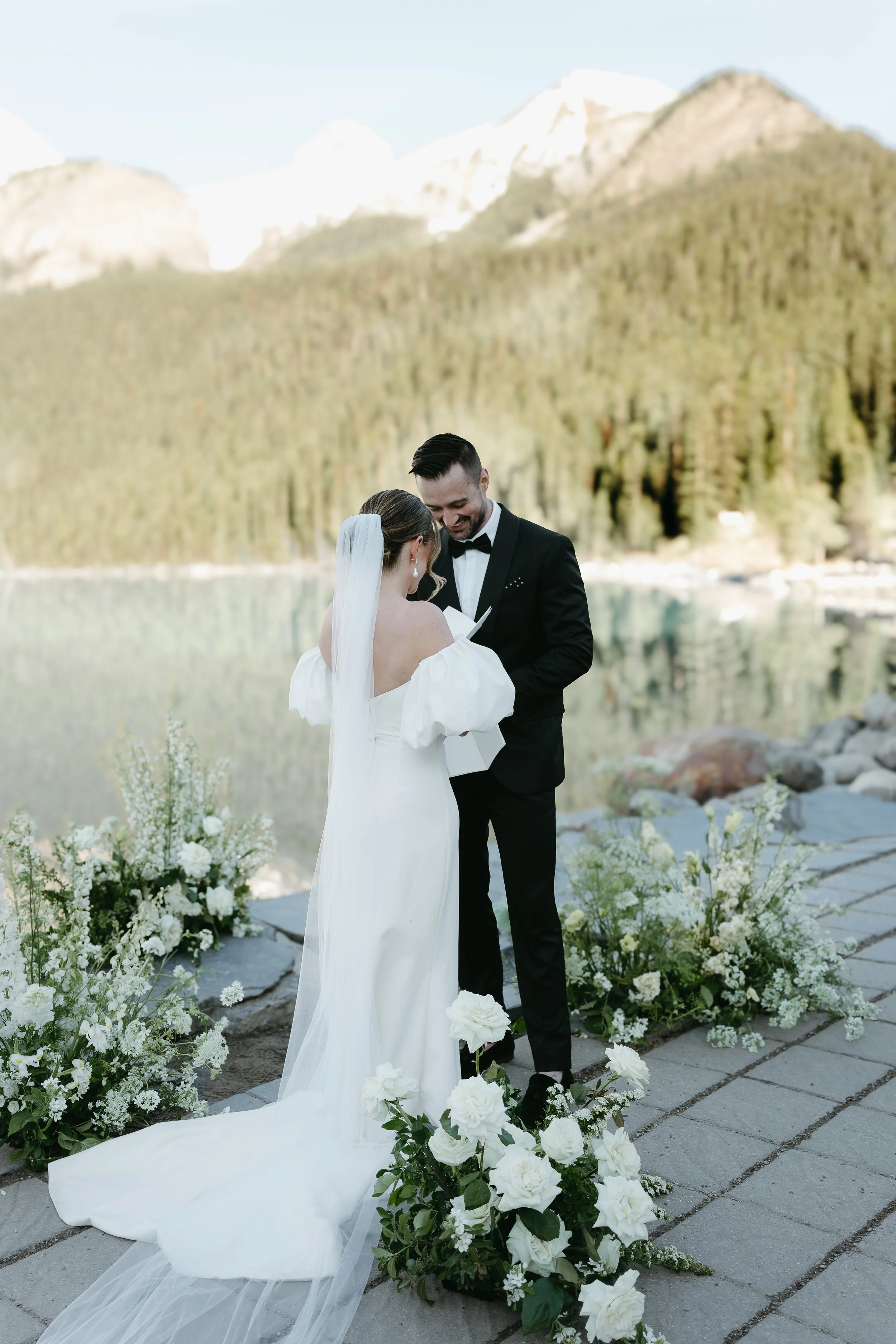 A bride and groom exchange vows on the edge of a lake surrounded by mountains. July 2025 Banff Elopement/Wedding Session with KC Photography