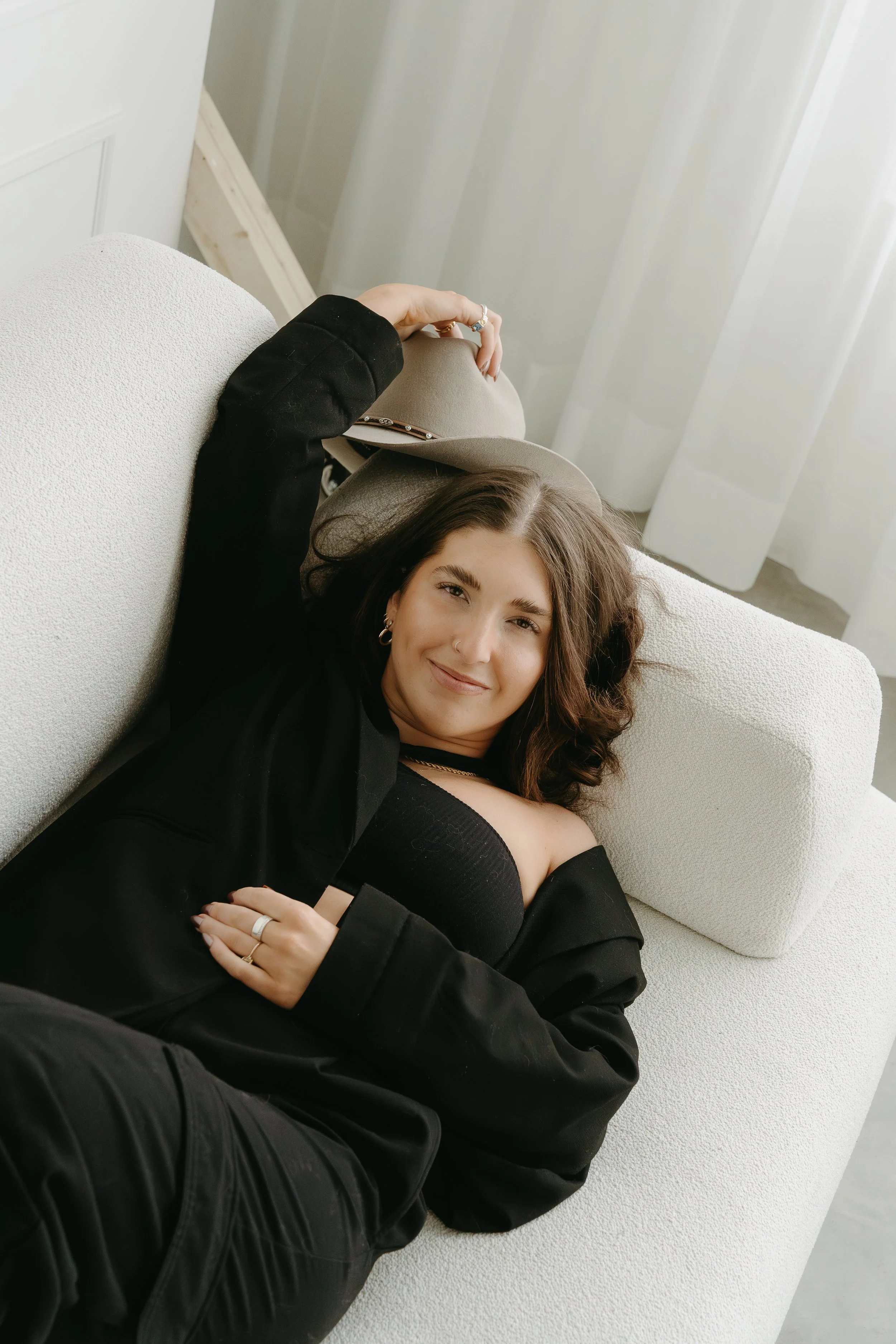A woman lays on a couch, holding her cowboy hat over the arm and smiling as she looks up at the camera. Calgary Studio Branding Session with KC Photography