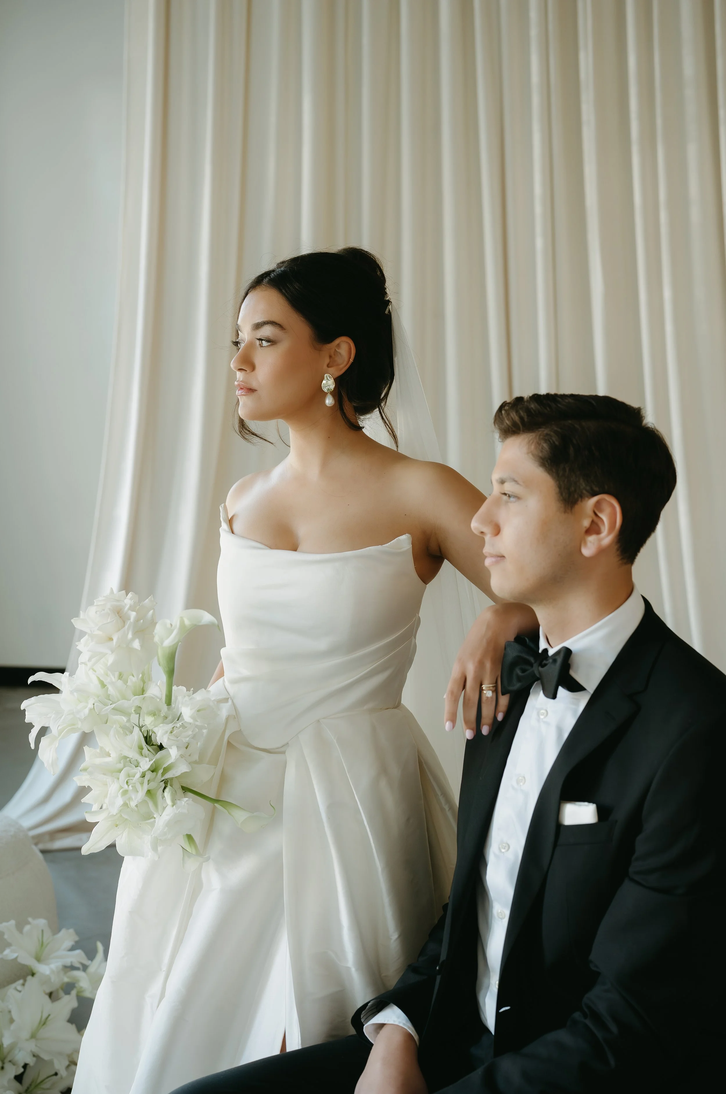 A groom sits in a chair while his bride stands beside him and they look into the distance in their empty reception. July 2025 Calgary Elopement/Wedding Editorial Session with KC Photography