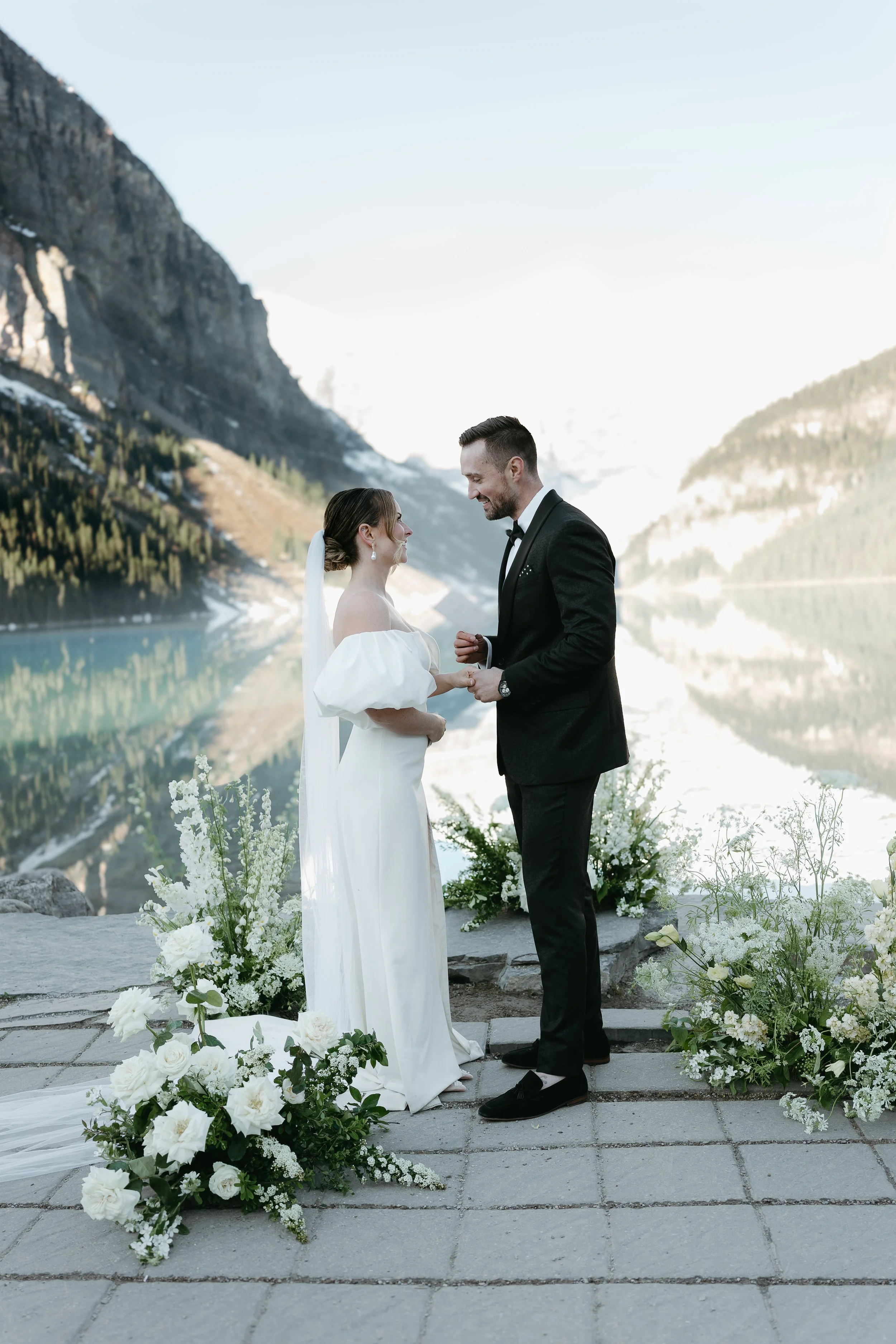 A bride and groom look at one another while holding each other's hand on the edge of a lake surrounded by bouquets of flowers and mountains.  August 2025 Canmore/Banff Elopement/Wedding Editorial Session with KC Photography