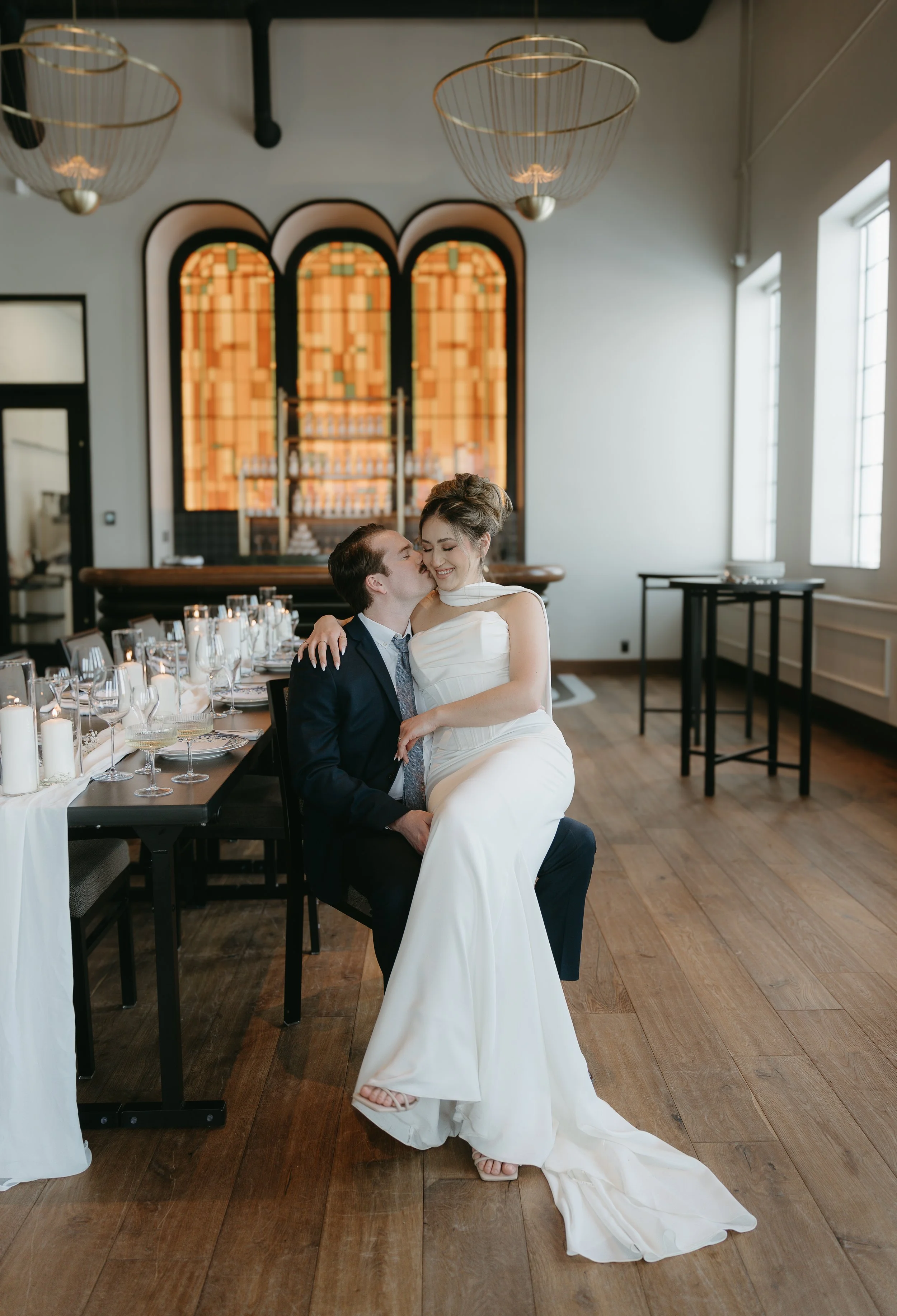 A bride sits on the lap of her groom in the empty reception while he kisses her on the cheek and she giggles. Calgary Elopement/Wedding Session with KC Photography