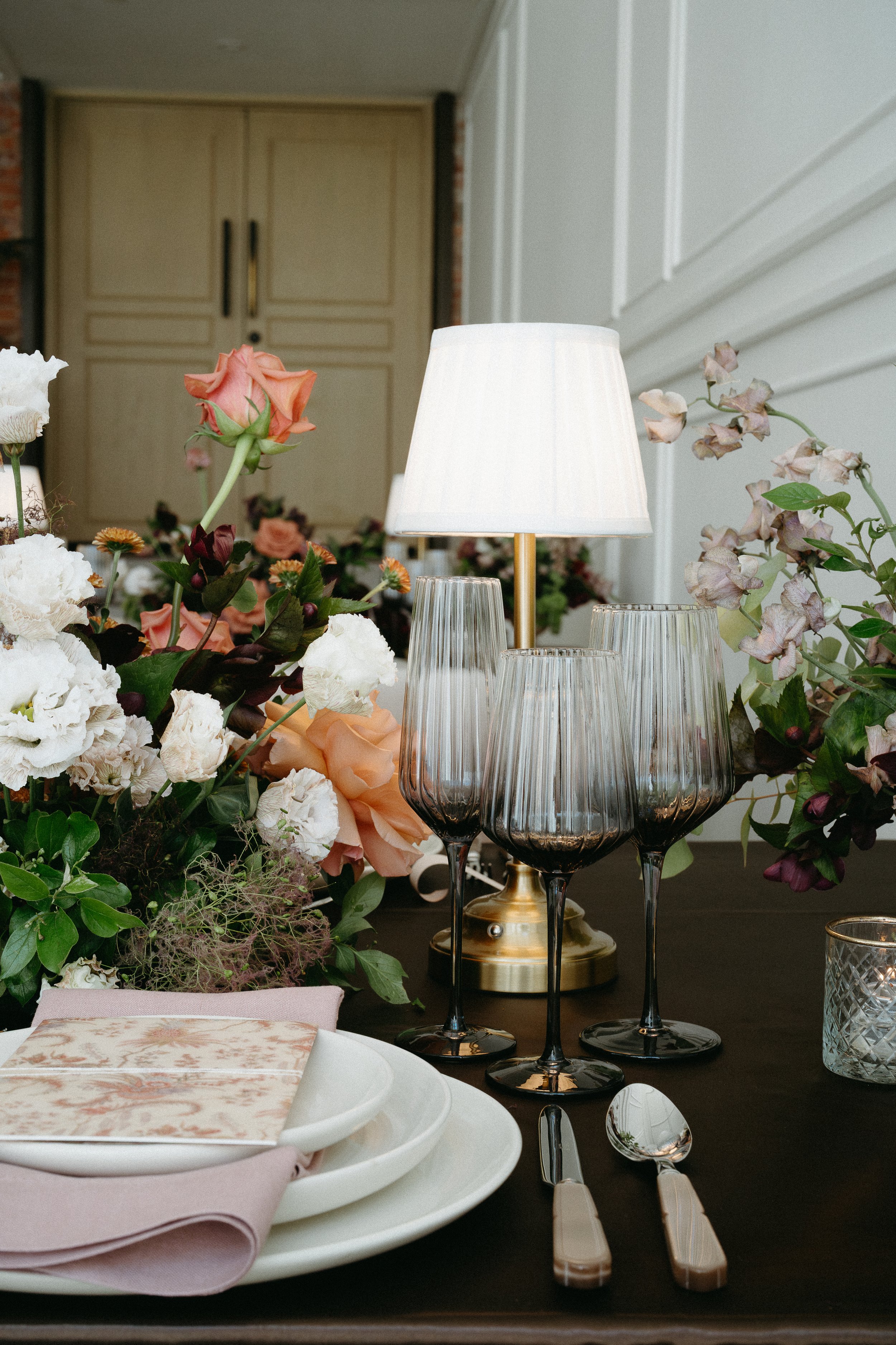 A tabletop at an empty wedding reception covered with table arrangements, bouquets of flowers, lamps, and candles. July 2025 Calgary Elopement/Wedding Editorial Session with KC Photography