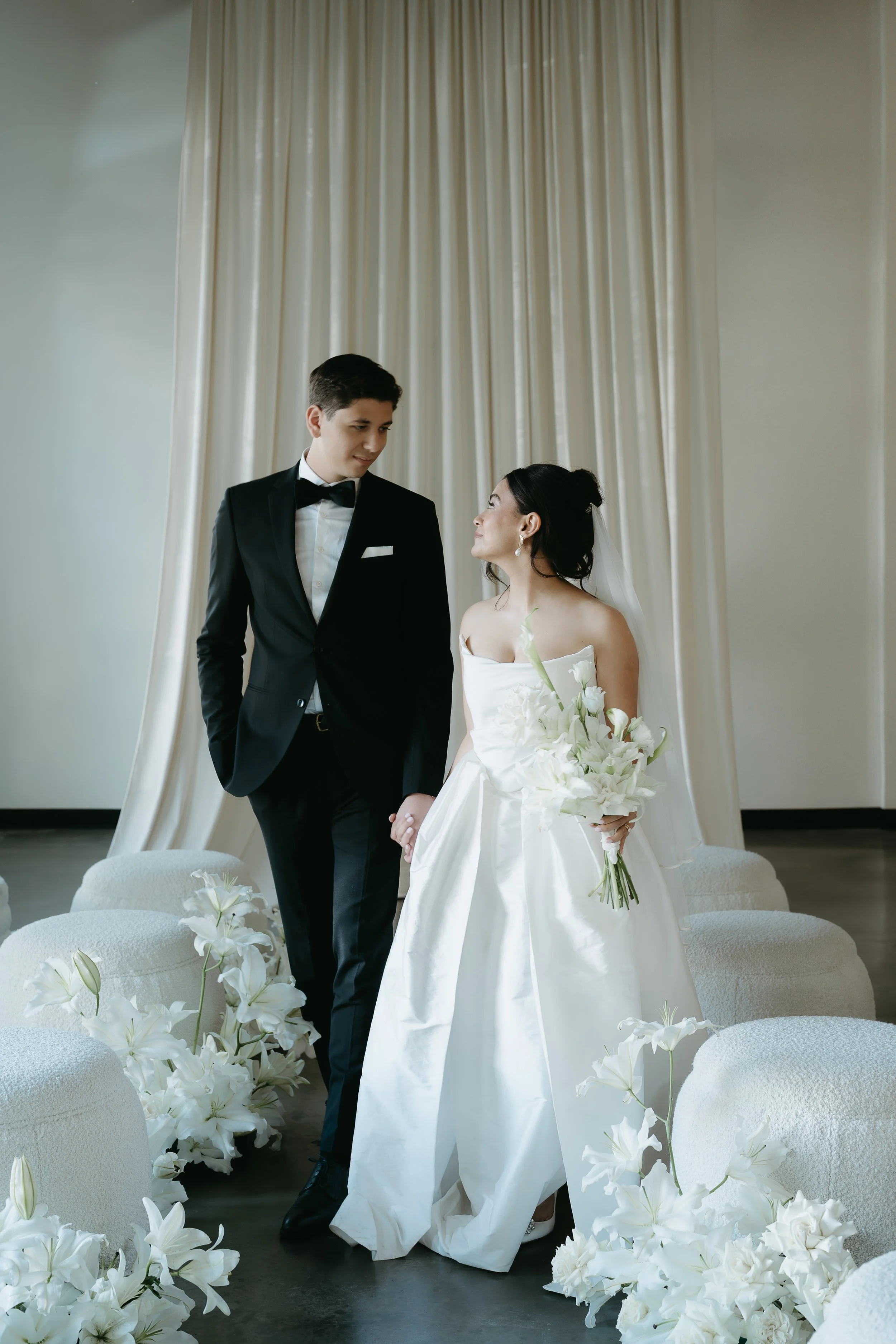 A bride and groom stand holding hands and looking at each other in an aisle of chairs and white lily's. July 2025 Calgary Elopement/Wedding Editorial Session with KC Photography
