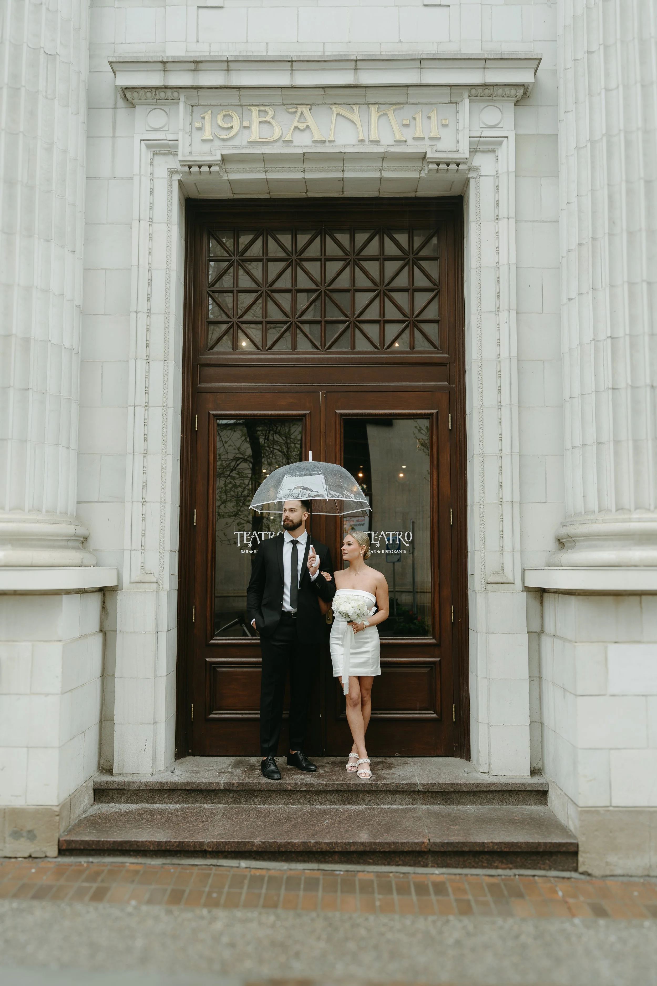 A groom shares an umbrella with his bride at the doors of Teatro in Downtown Calgary. Downtown Calgary Elopement/Wedding Session with KC Photography