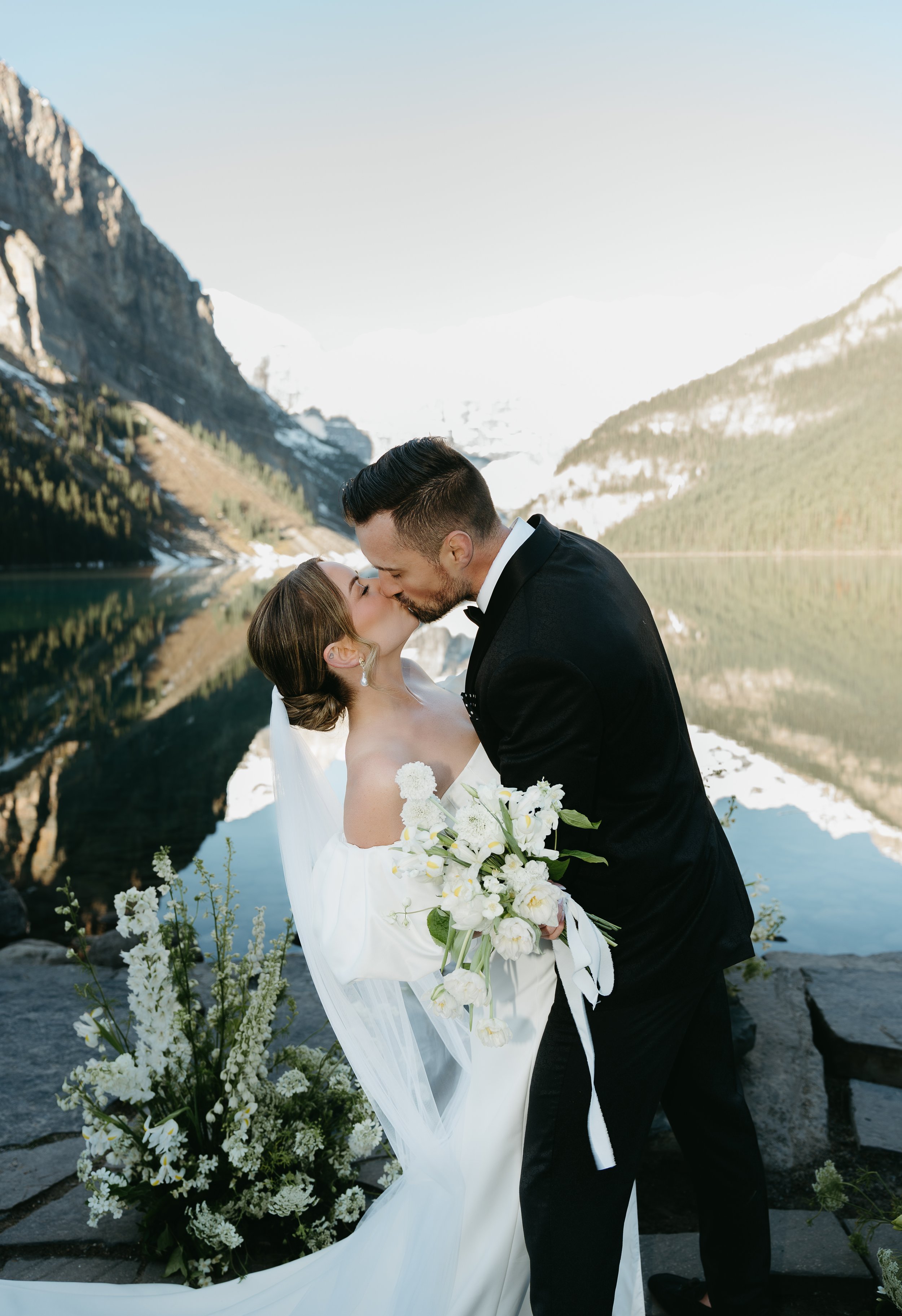 A bride and groom share a kiss on the edge of a lake surrounded by mountains. August 2025 Canmore/Banff Elopement/Wedding Editorial Session with KC Photography