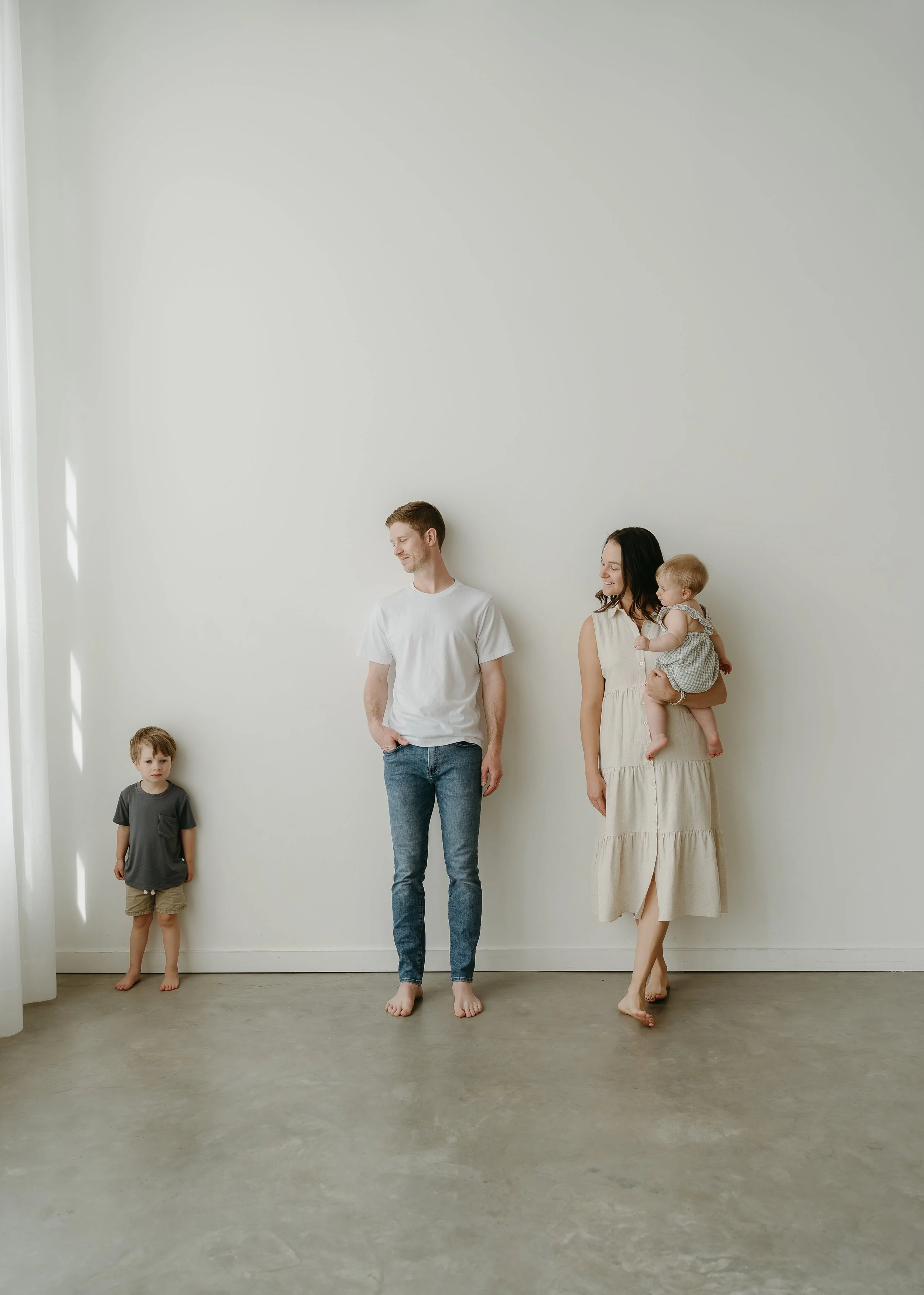 A mother holds her infant while leaning against the white wall of a studio beside her husband who all are looking beside them at their toddler standing beside them looking at the camera. Calgary Lifestyle/Family Studio Session with KC Photography