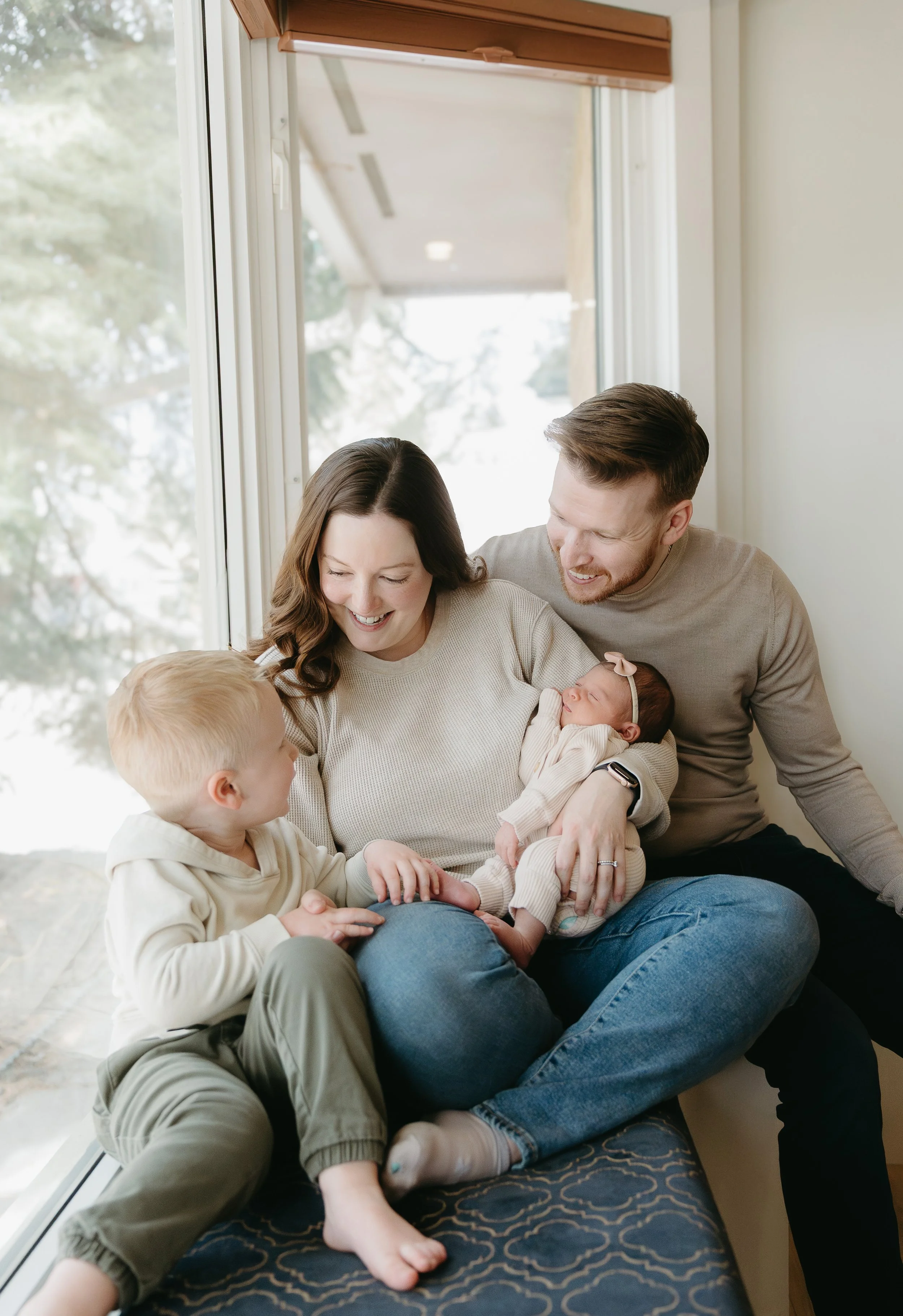 A mother holds her newborn baby while sitting beside her husband in the window sill as they look down at their toddler sat next to them.  Calgary Lifestyle/Family/Newborn Home Session with KC Photography