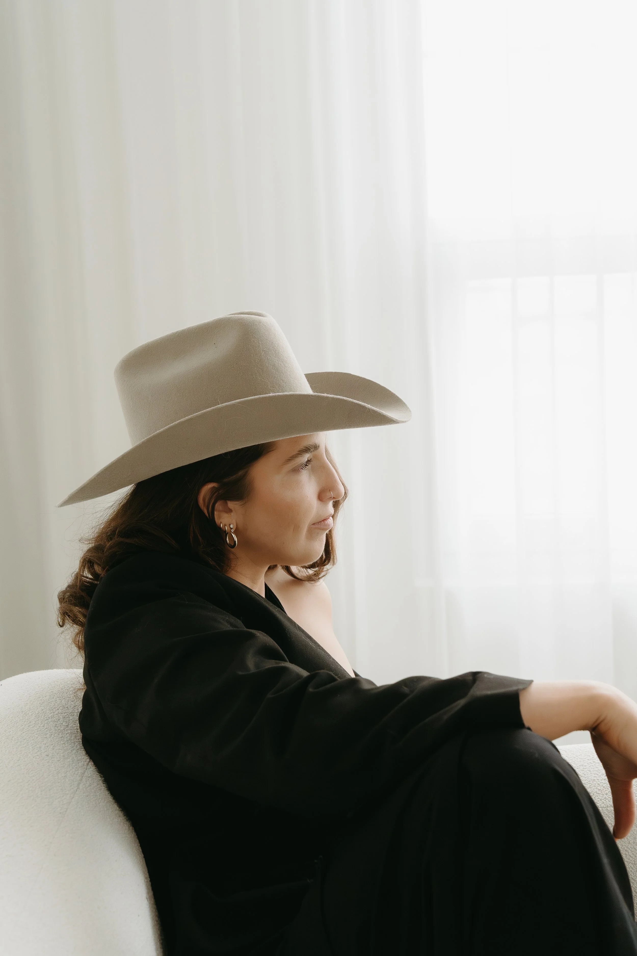 A woman's [profile is caught as she sits on a couch and looks into the distance wearing a cowboy hat. Calgary Studio Branding Session with KC Photography