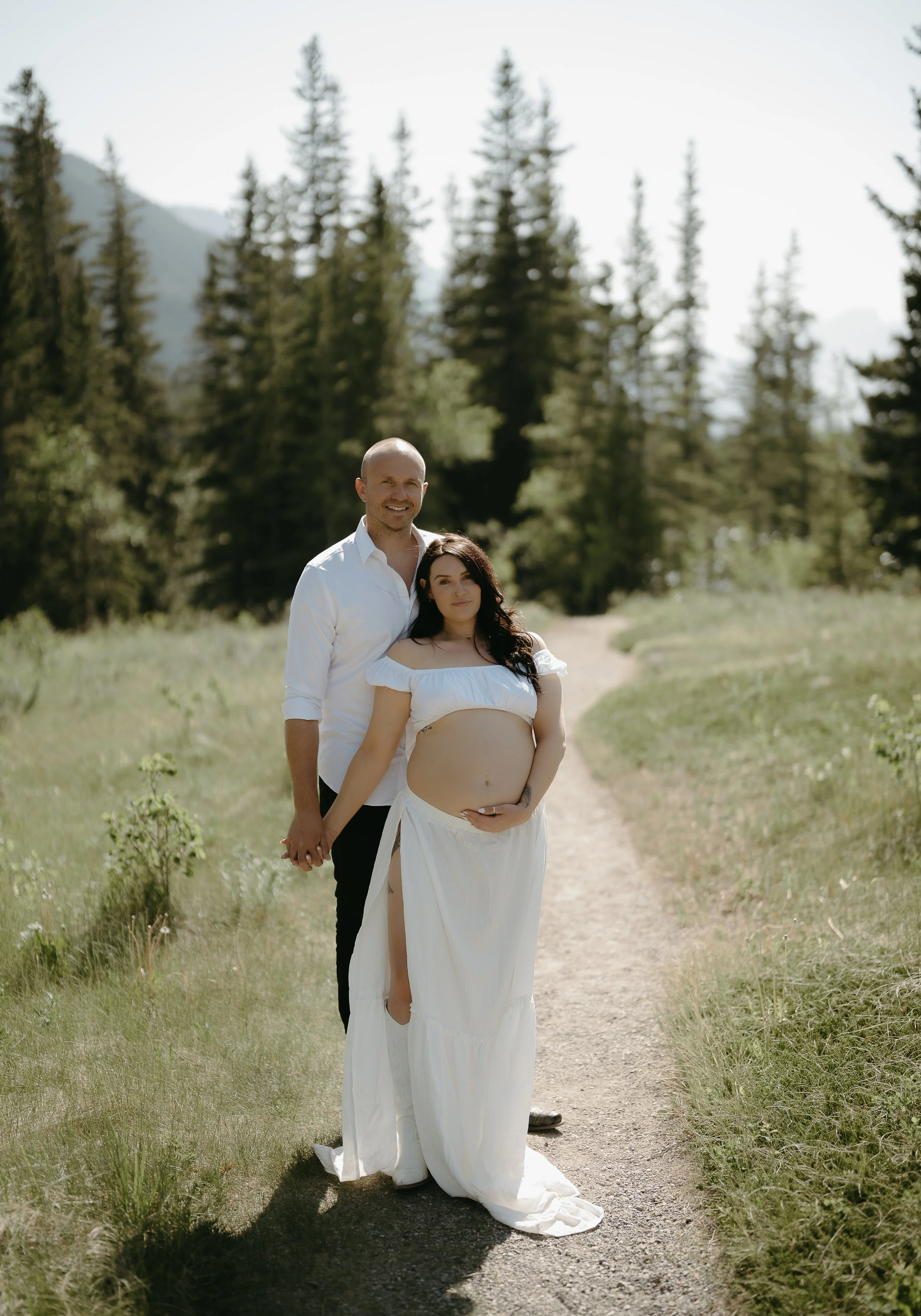 A pregnant women holds the hand of her husband as he holds her from behind on a Kananaskis path in a field. Kananaskis Lifestyle/Family/Couple/Pregnancy/Maternity Session with KC Photography