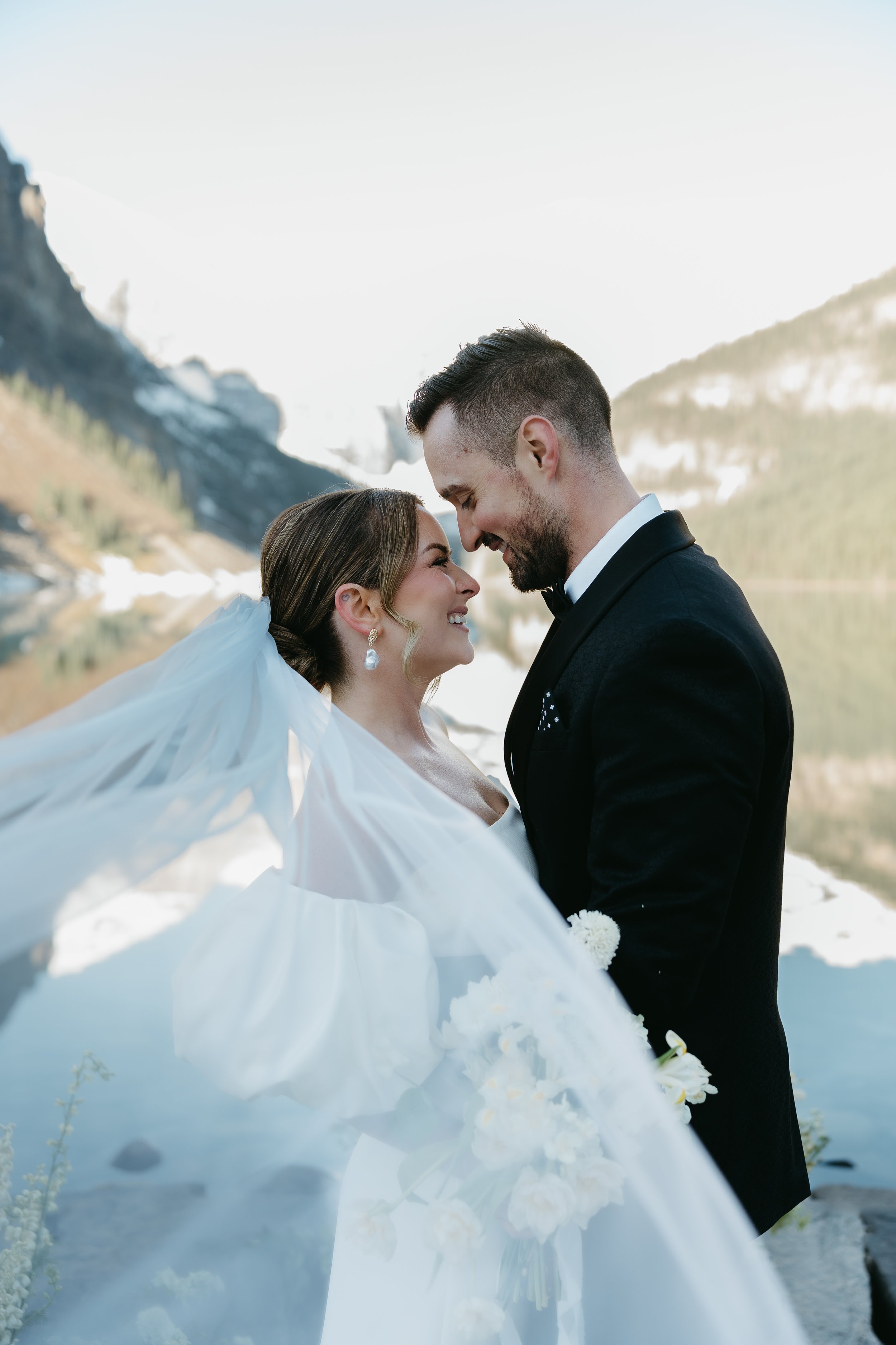 A bride and groom hold one another closely and smile together as the bride's veil flows in the wind on the edge of a lake surrounded by mountains. August 2025 Canmore/Banff Elopement/Wedding Editorial Session with KC Photography