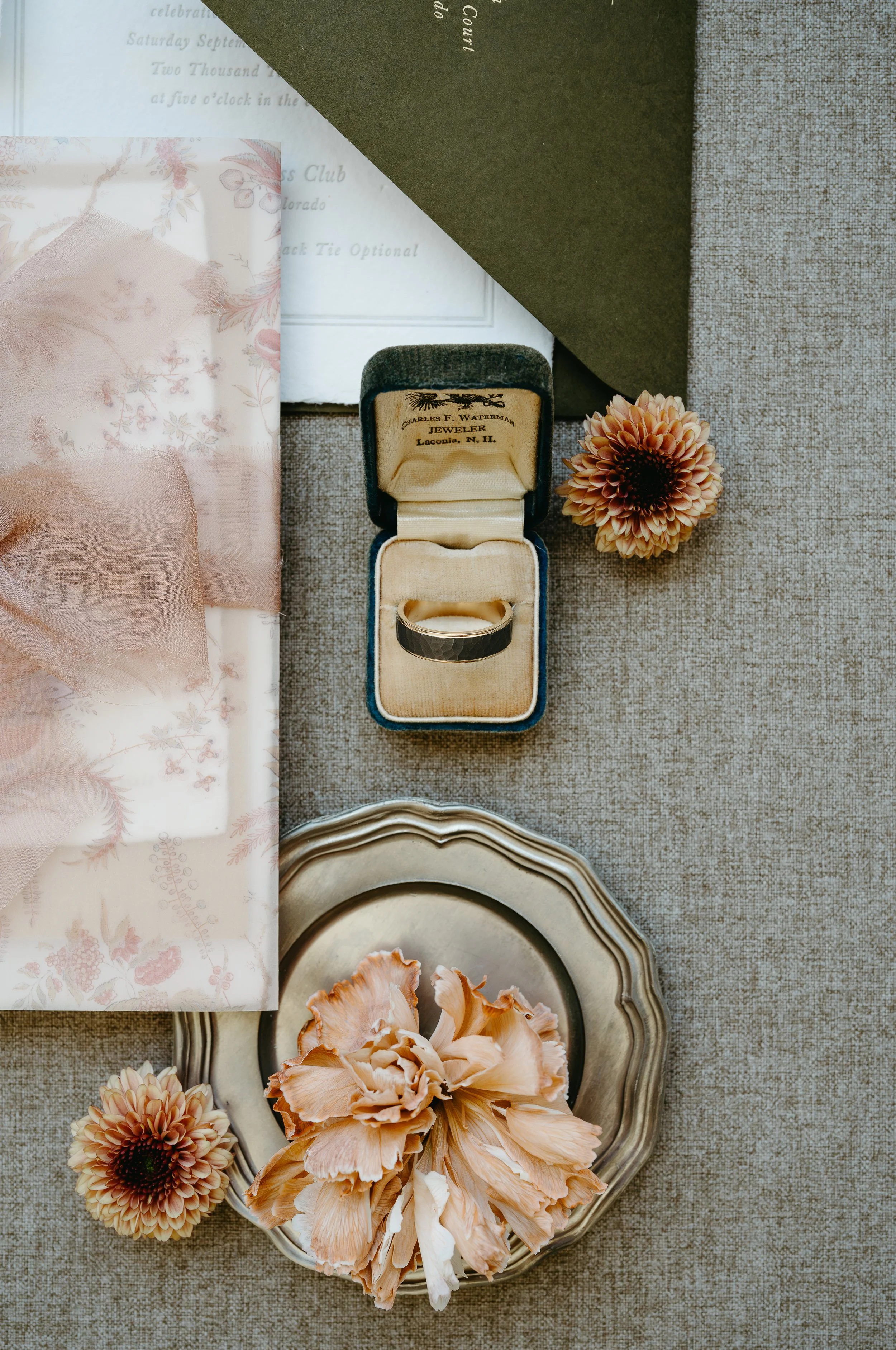 A wedding band is placed on a grey surface surrounded by flowers and wedding invitations. July 2025 Calgary Elopement/Wedding Editorial Session with KC Photography