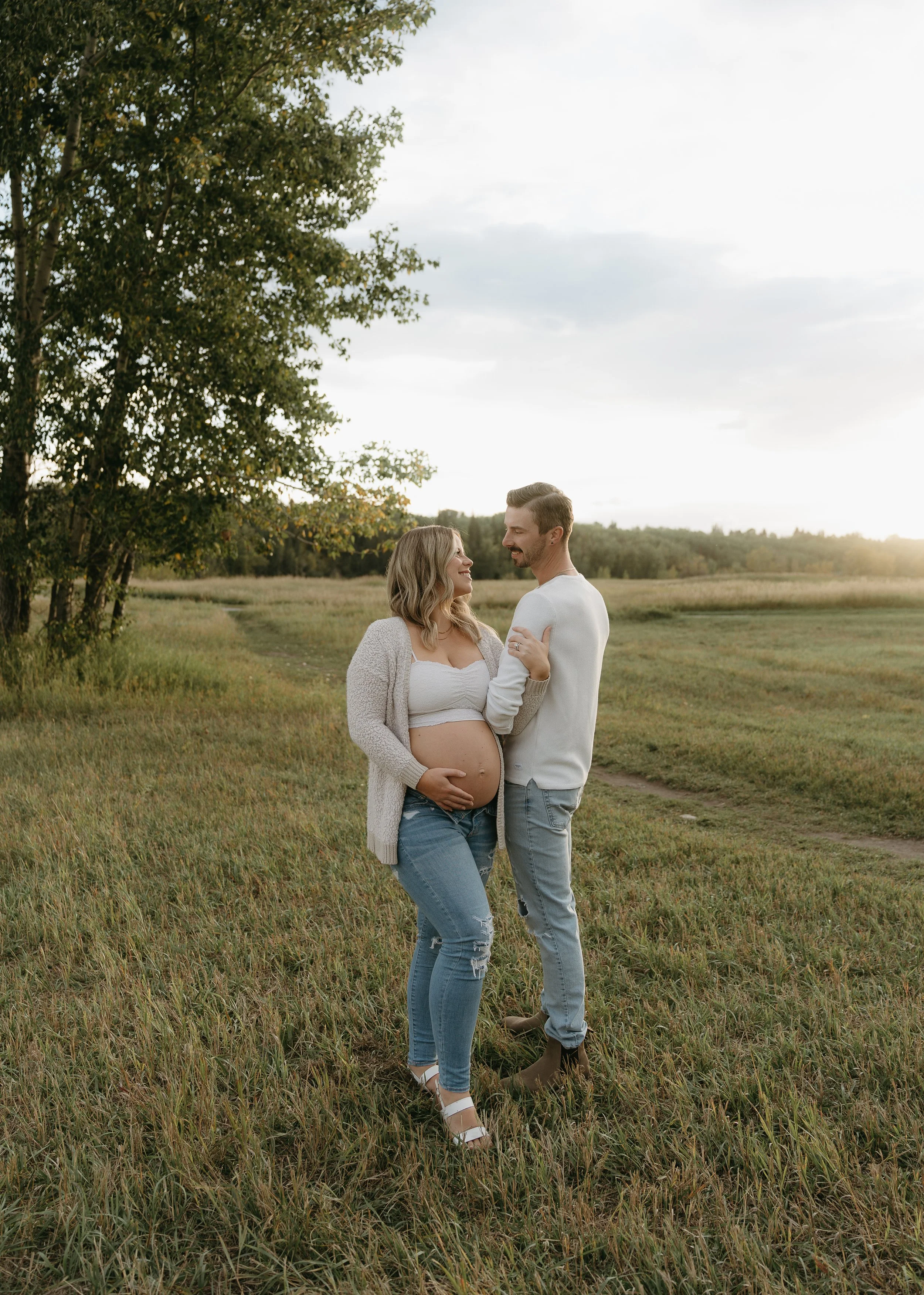 A woman wraps her arm around her husband's as they smile at one another while she holds her pregnant belly in a field. Kananaskis Lifestyle/Couple/Pregnancy/Maternity Session with KC Photography
