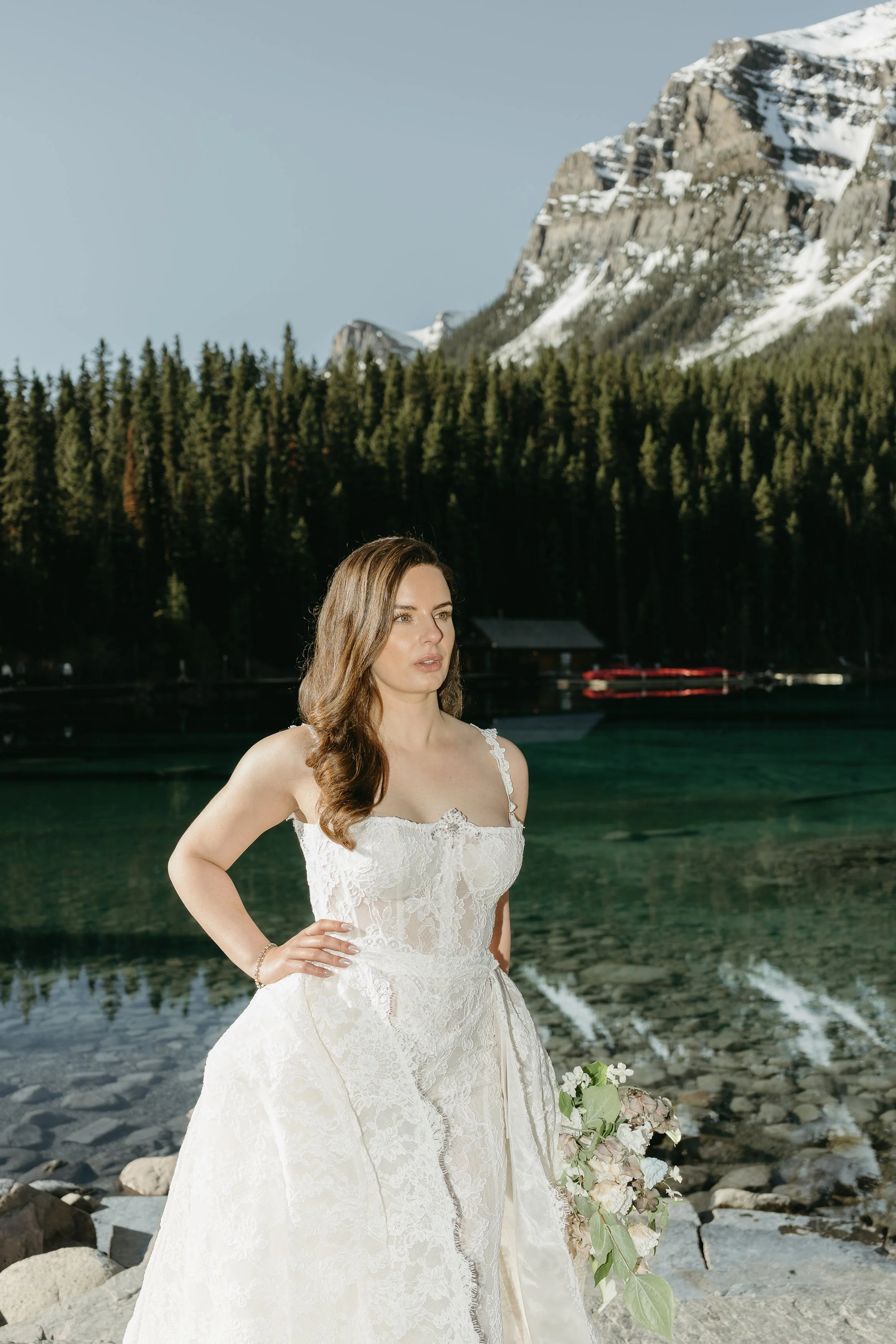 A bride stands on the edge of Lake Louise with a mountain pictured in the background. June 2025 Lake Louise Elopement/Wedding Editorial Session with KC Photography